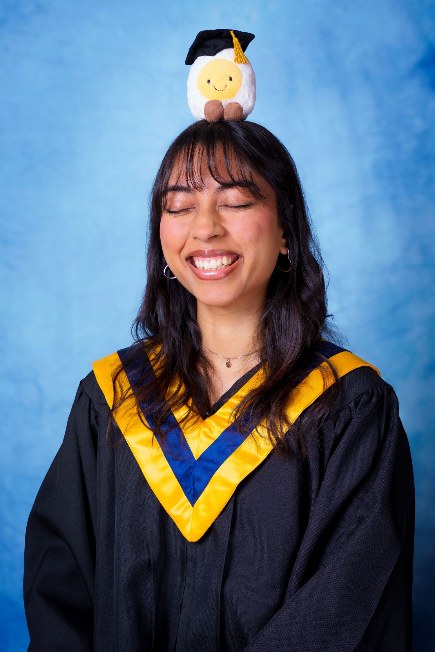 Graduate in academic regalia smiles joyfully against blue background.
