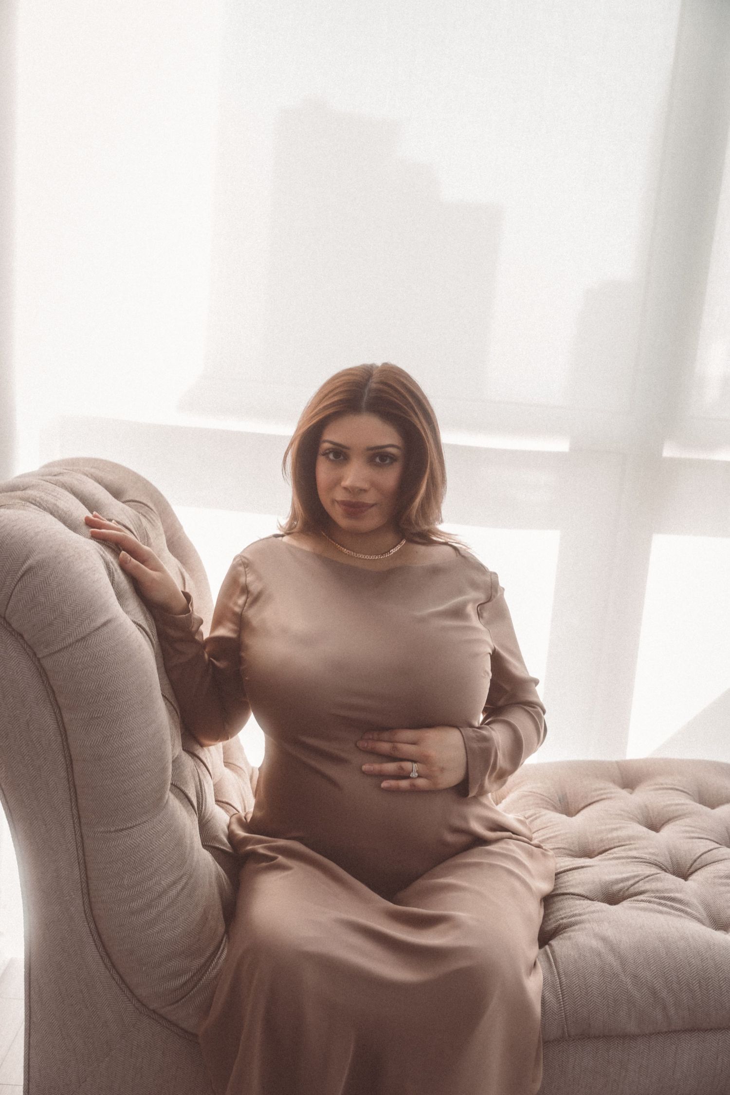 A person in a beige long-sleeved dress sits on a tufted gray chaise lounge near a bright window.