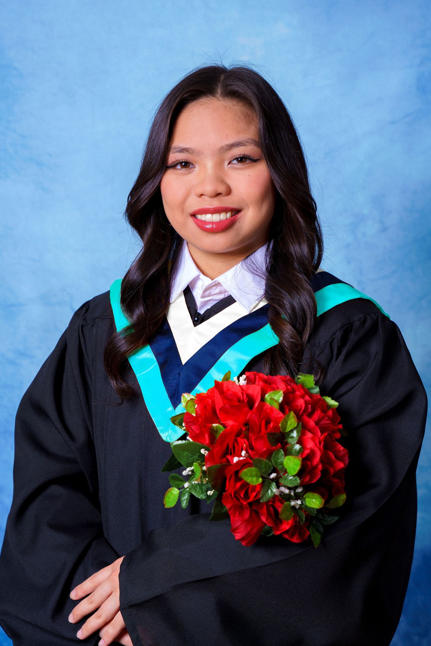 Graduate wearing academic regalia poses with vibrant red bouquet against blue background.