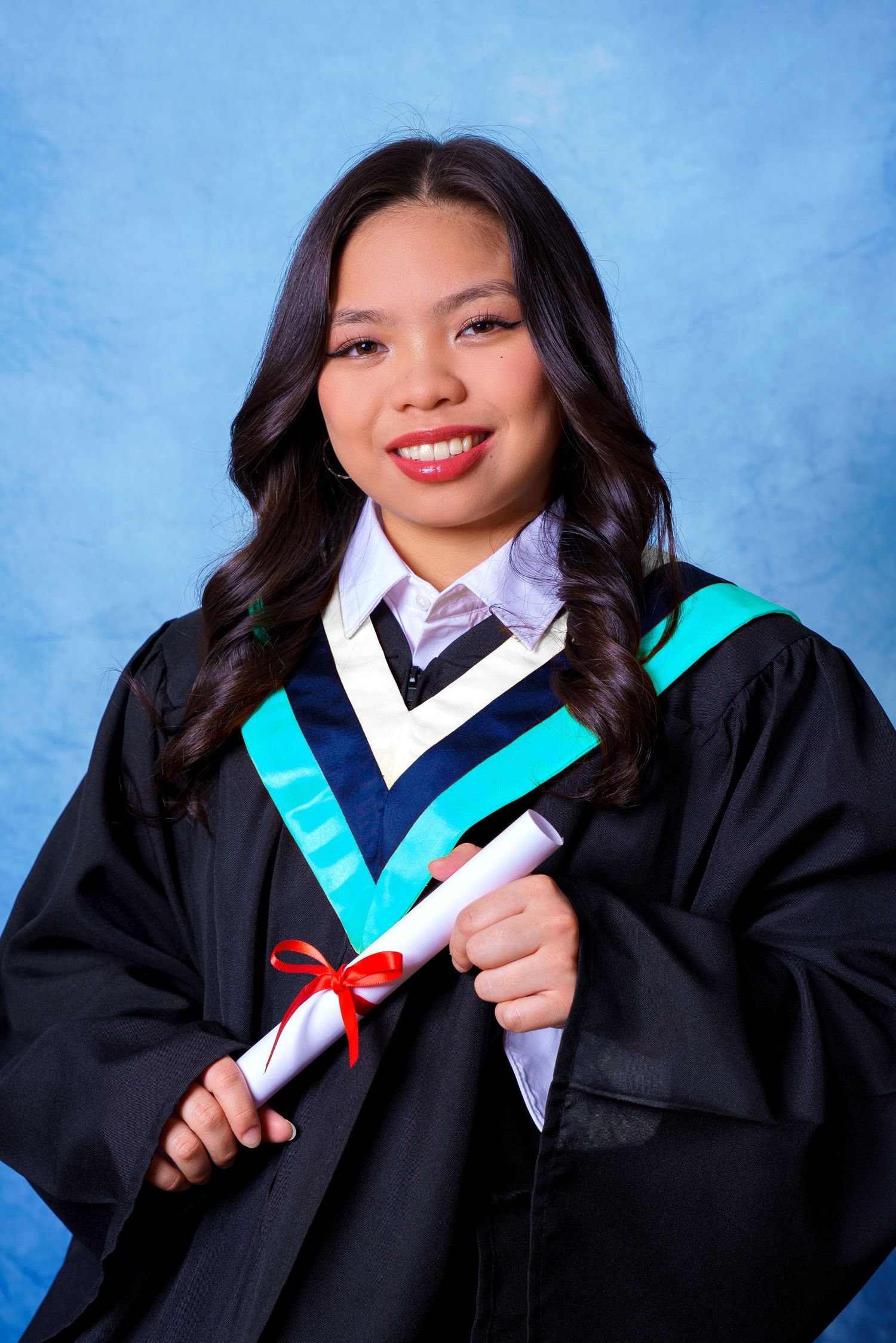 Graduate in black academic regalia with turquoise trim holds diploma against blue backdrop.