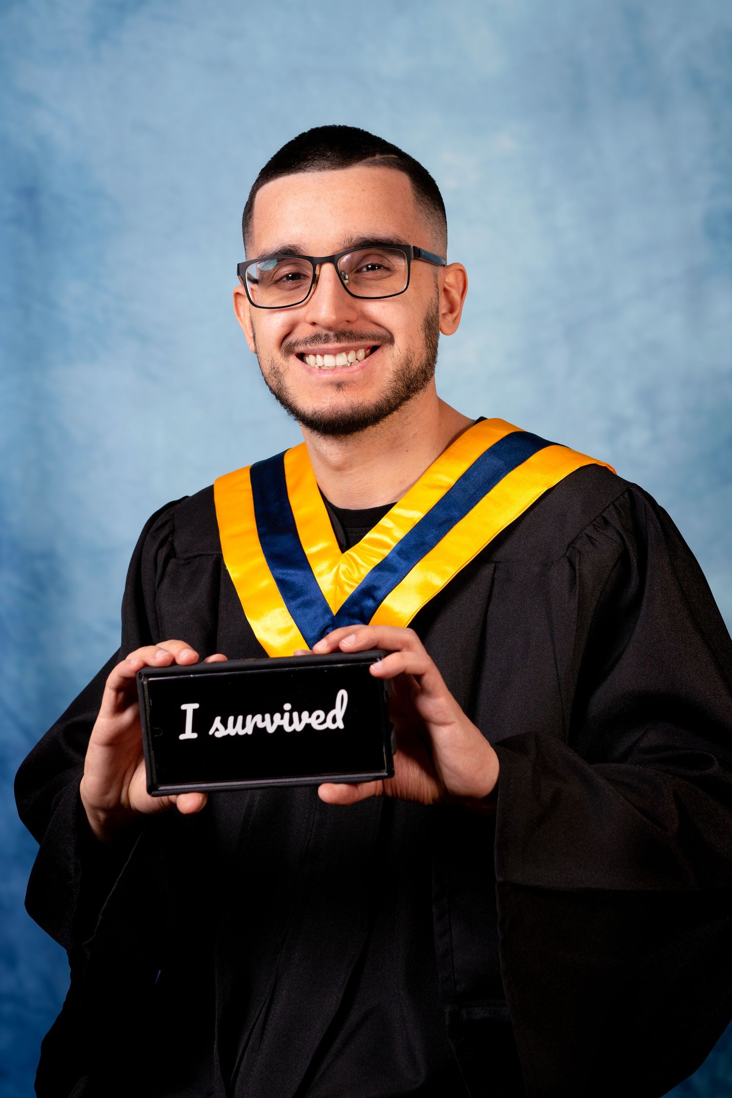 Graduate in black academic regalia with gold and blue chevrons holds phone displaying 'I survived' text against blue backdrop.