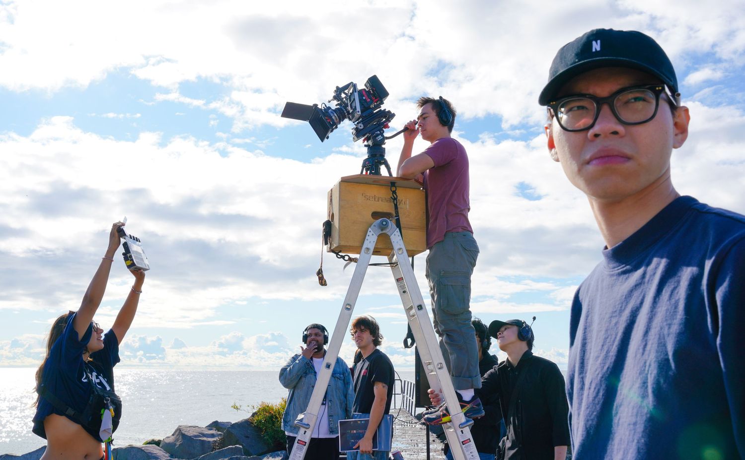 Film production crew setting up camera equipment against a bright sky backdrop.