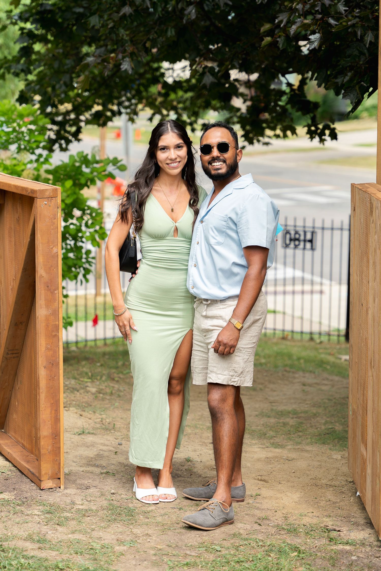 A couple poses together outdoors, with her wearing a mint green dress and him in a white shirt and khaki shorts.
