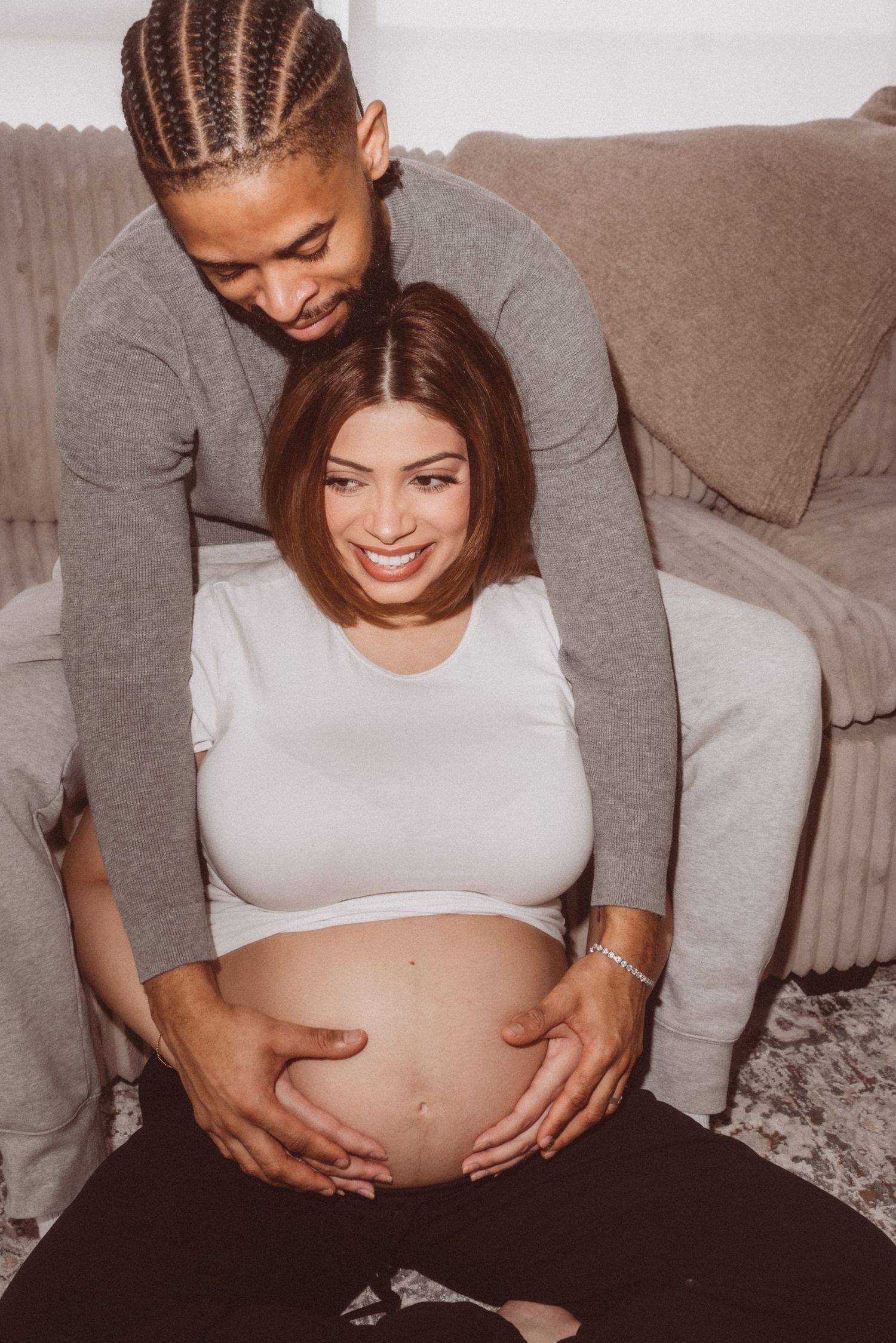 A couple shares an intimate moment as they embrace while showing off a pregnant belly in a warm-toned photograph.