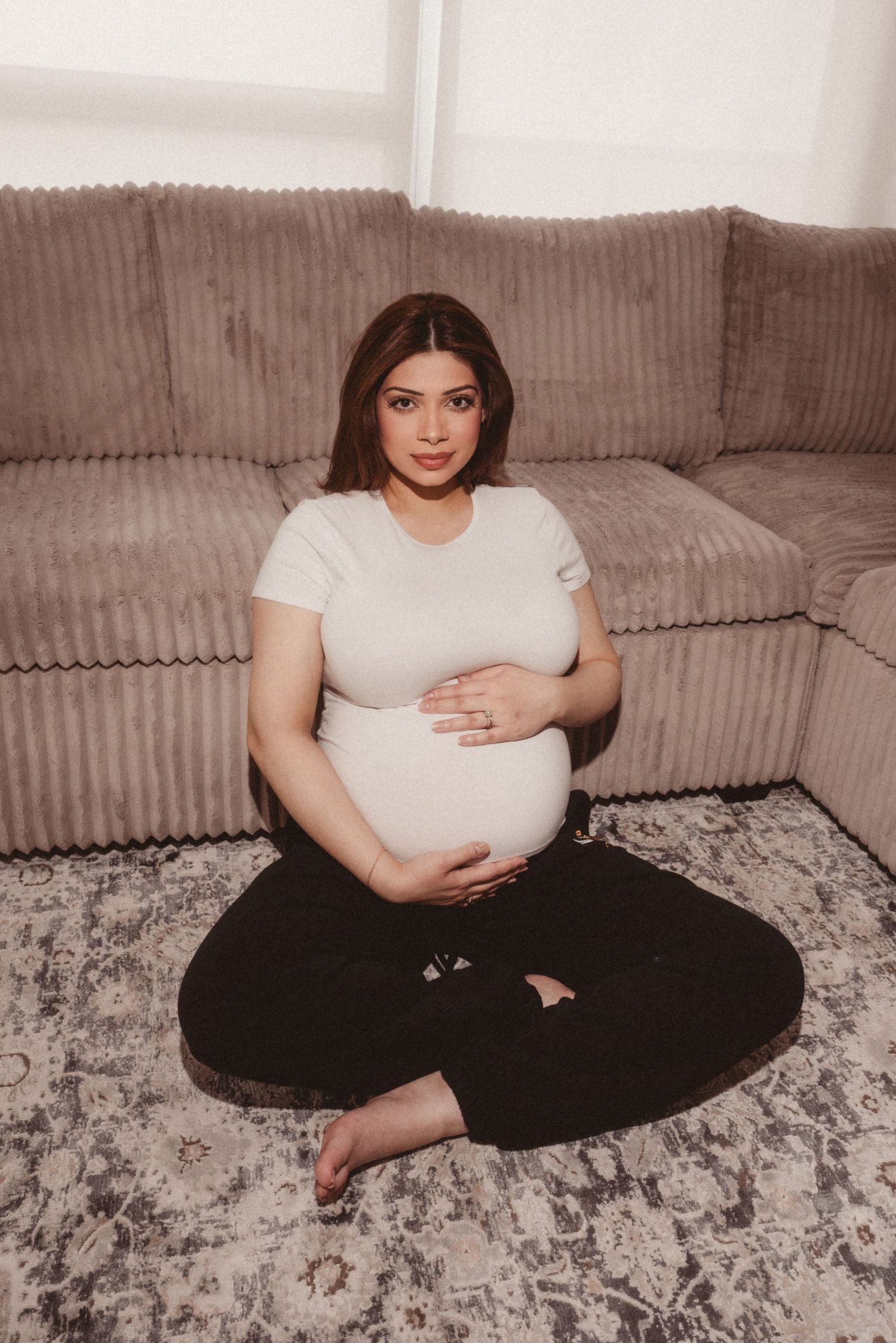 A pregnant woman wearing a white t-shirt and black pants sits cross-legged on a gray patterned rug by a beige sectional sofa.