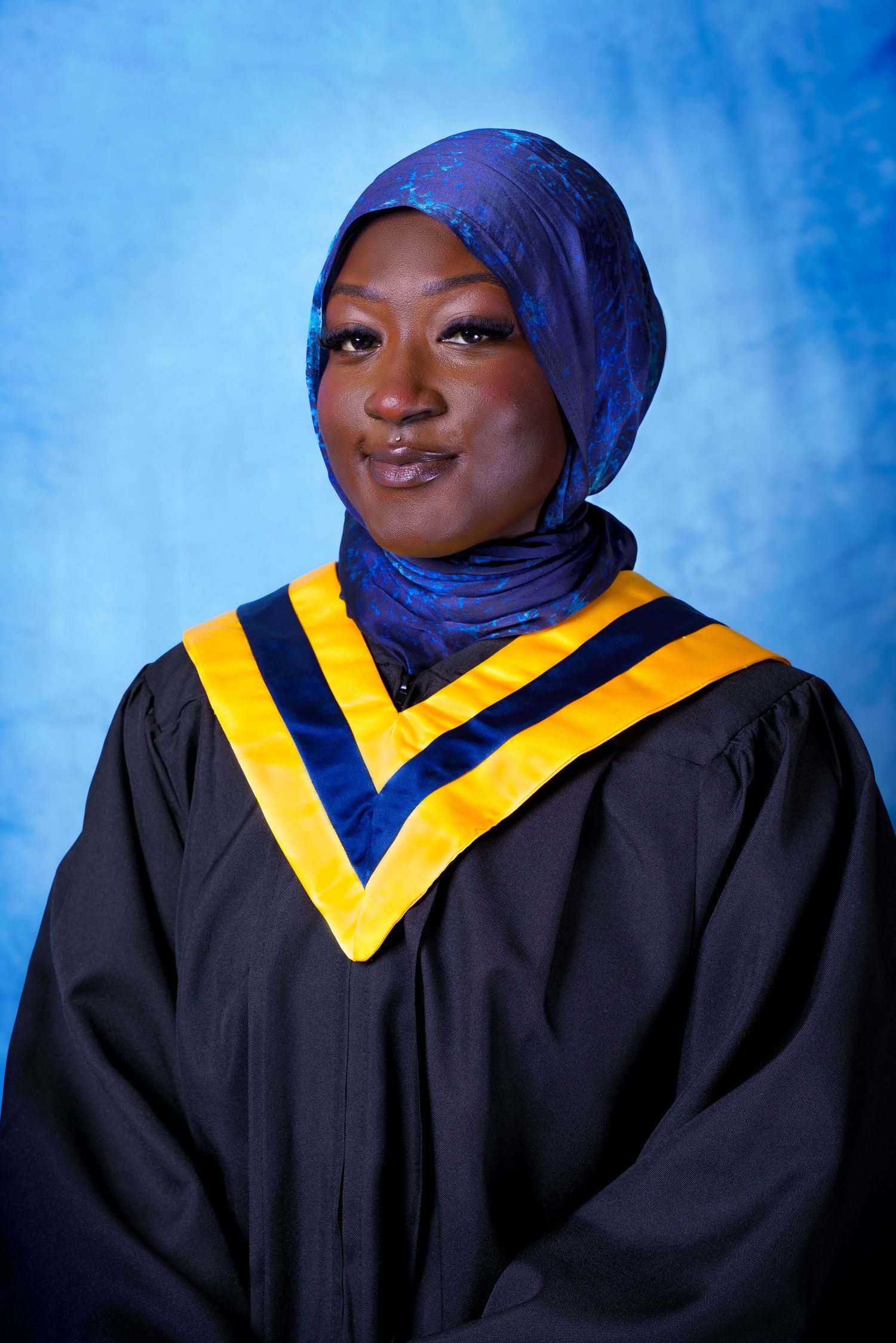 Graduate in blue hijab and black academic gown with gold trim poses against blue backdrop.