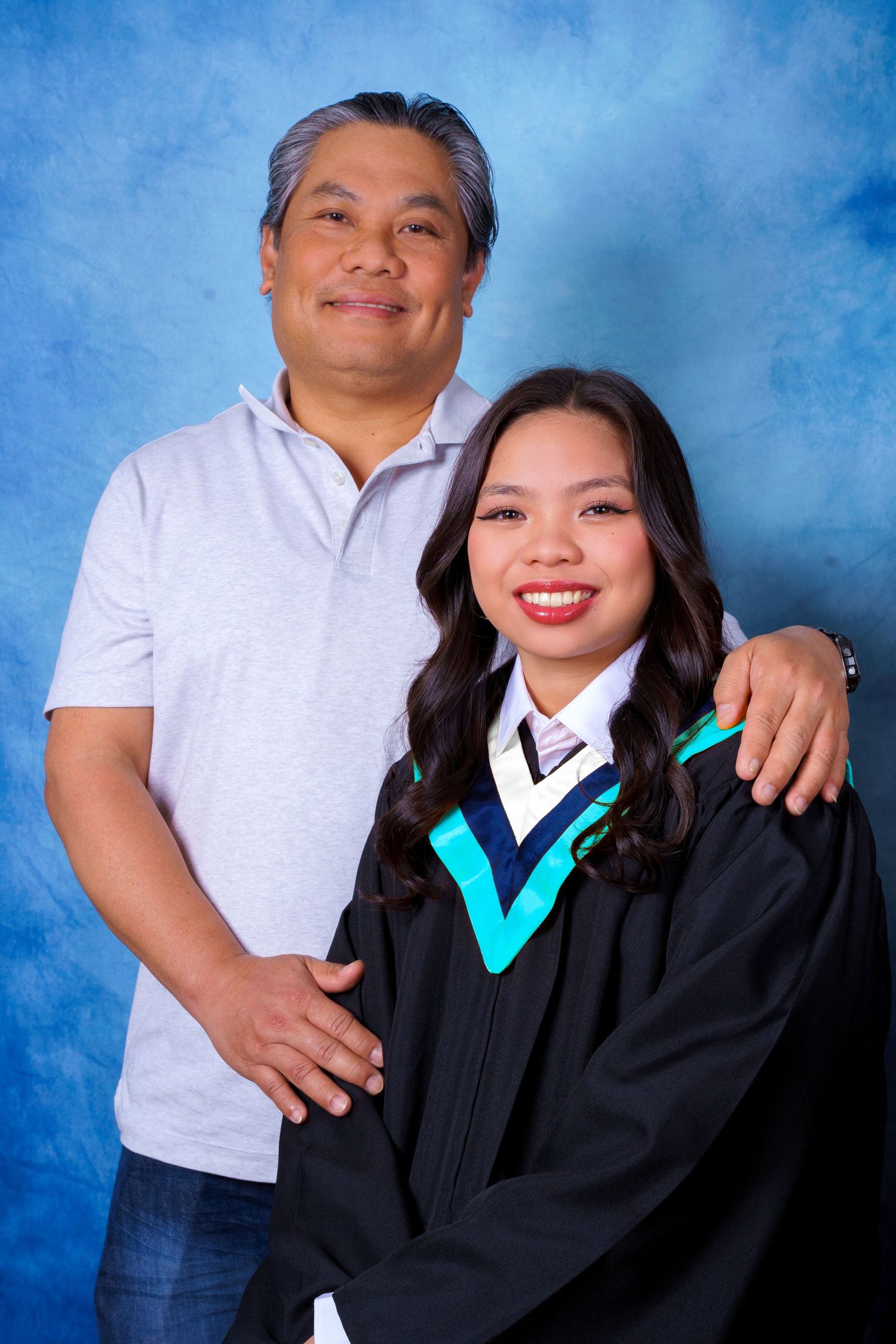 Graduation portrait featuring graduate in academic regalia with family member against blue backdrop.