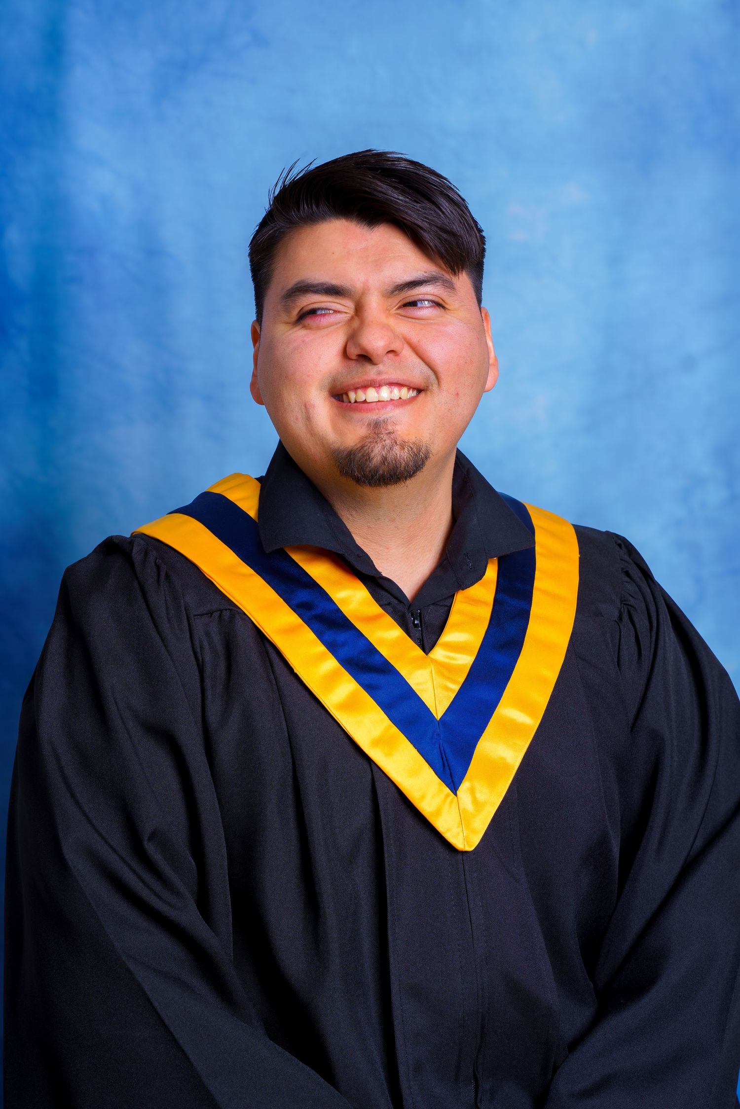 Graduate wearing black academic gown with gold and blue chevrons poses against light blue backdrop.