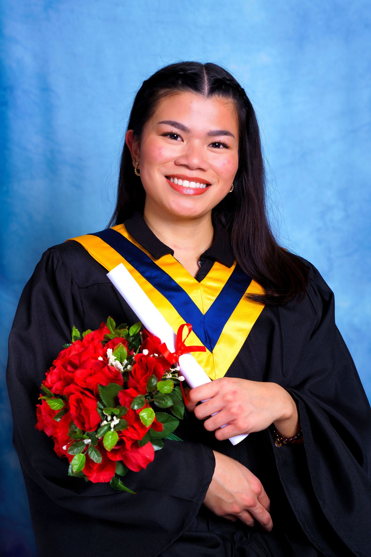 Graduate in black and gold academic regalia holds red floral bouquet for portrait.