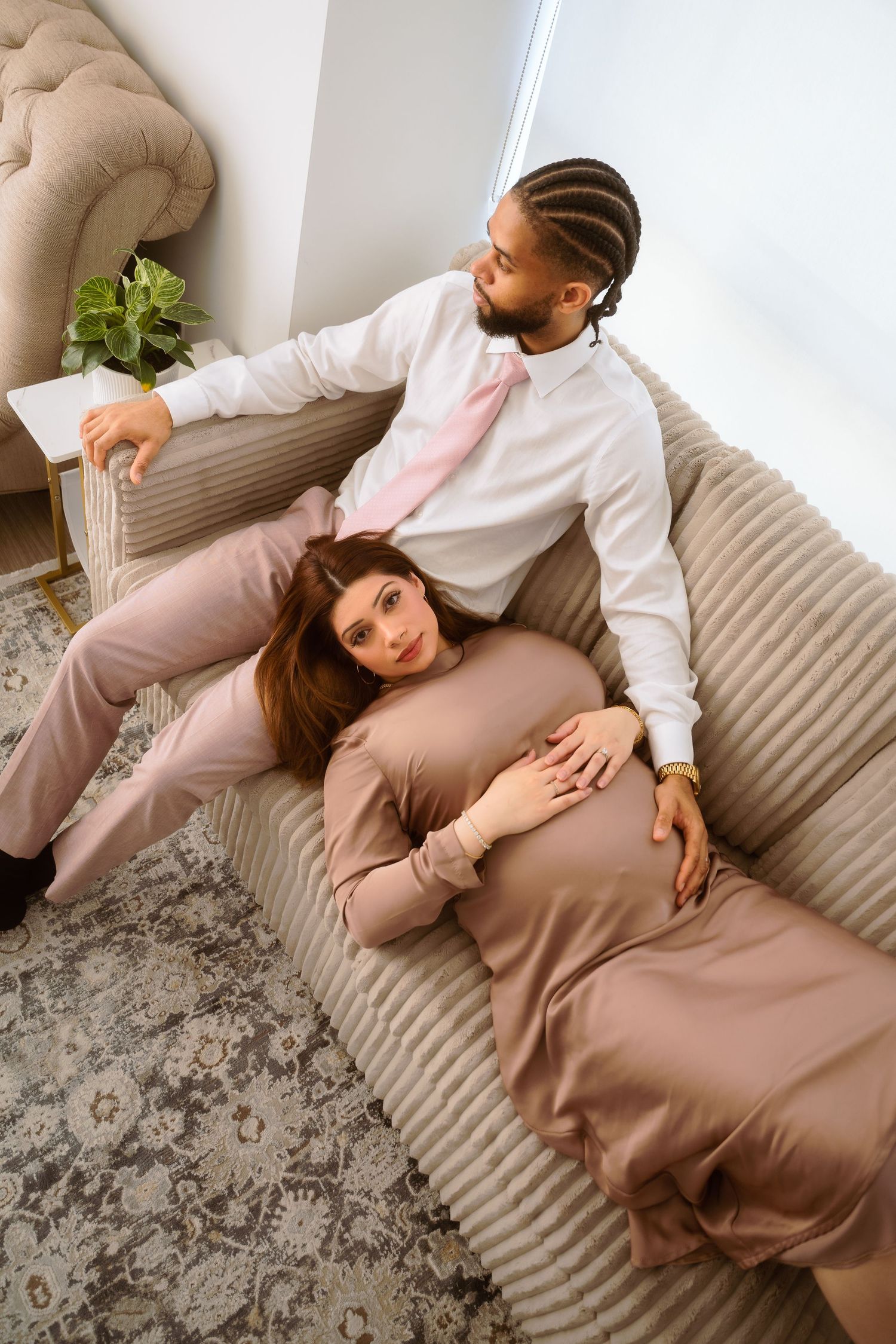 A couple lounges together on a beige couch in coordinating neutral outfits creating an intimate maternity photoshoot.