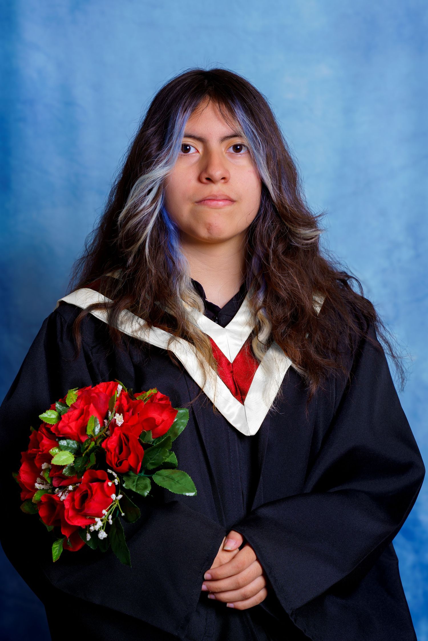 Graduate in academic regalia holds red roses while posing against blue background.