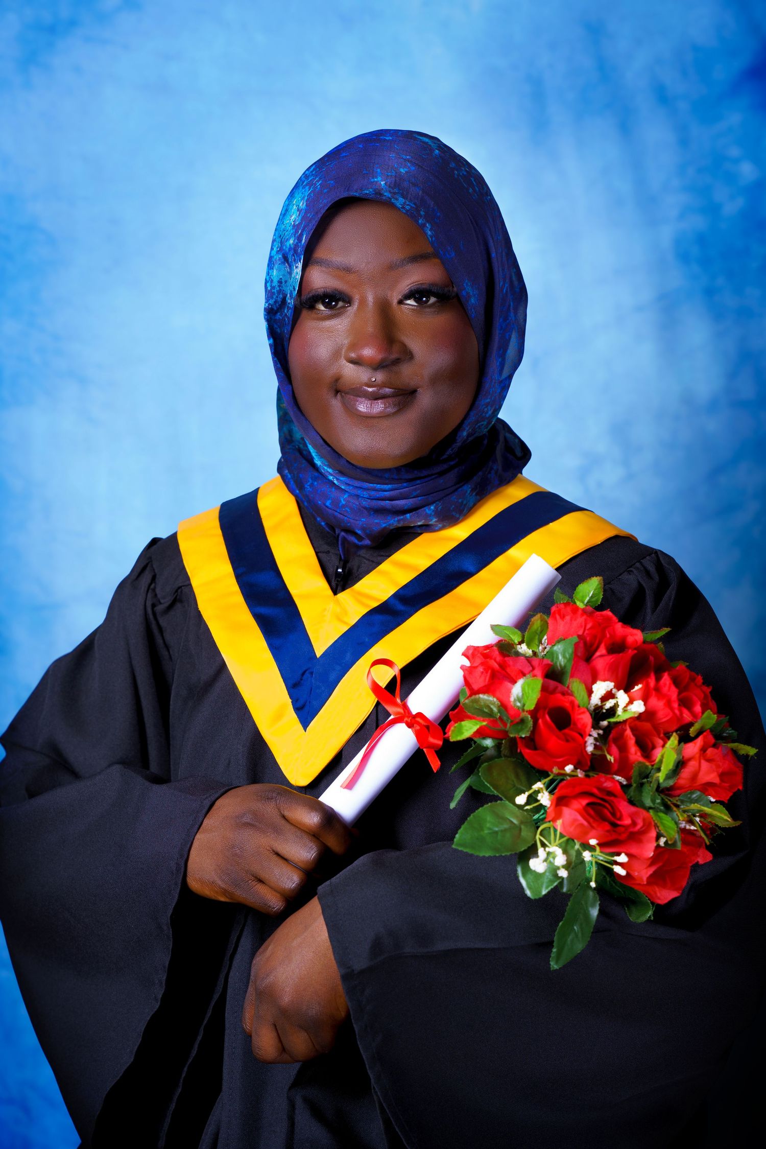 Graduate in hijab and academic regalia holds diploma and red roses against blue background.