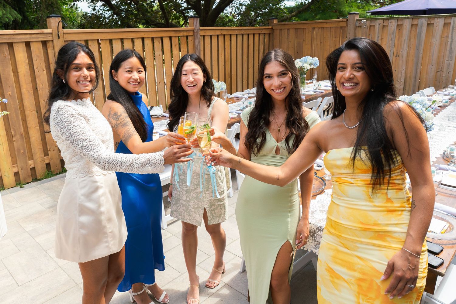 Five women in colorful dresses toast with champagne glasses at an outdoor celebration.
