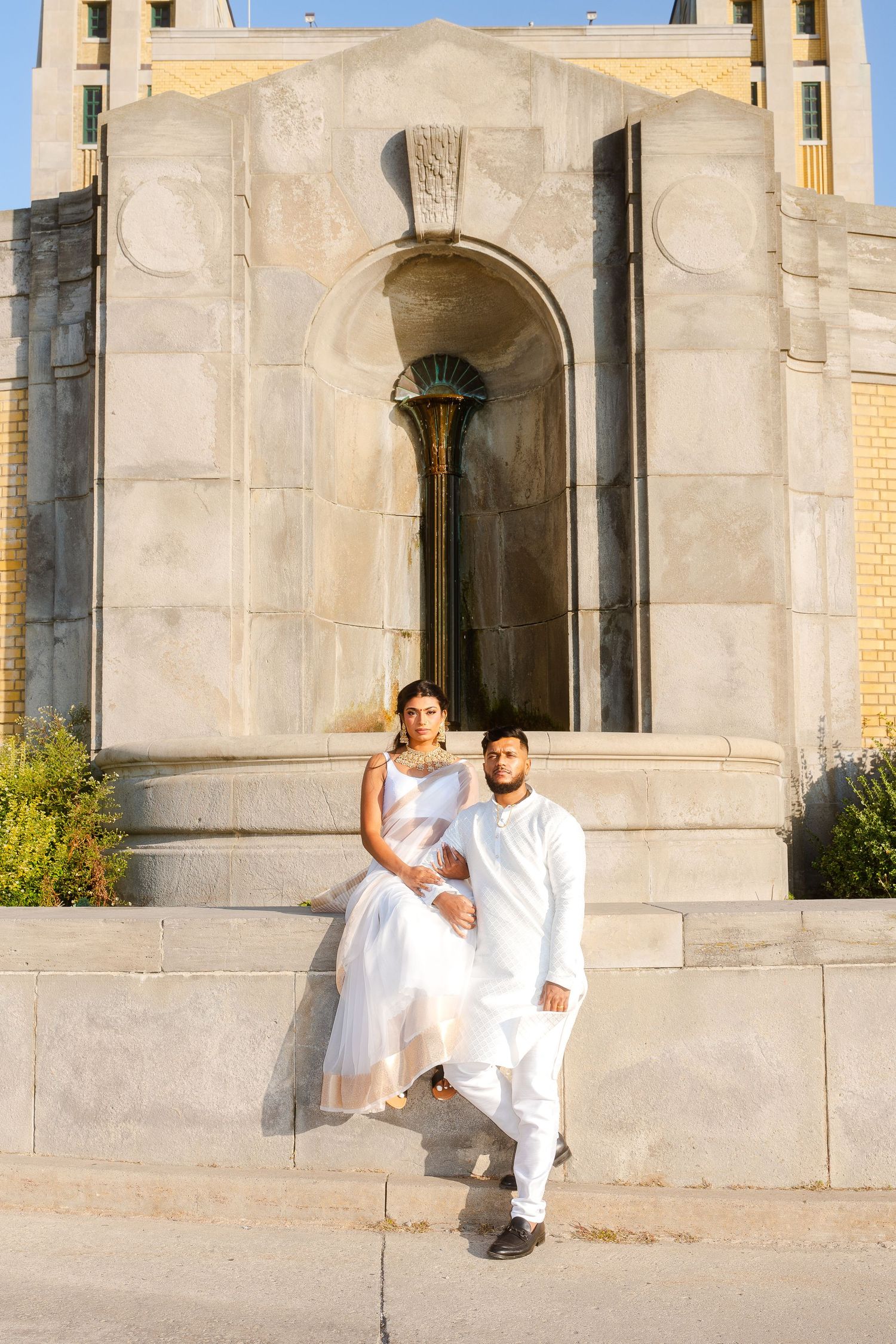 A couple in white outfits poses together against a stone church or cathedral exterior.