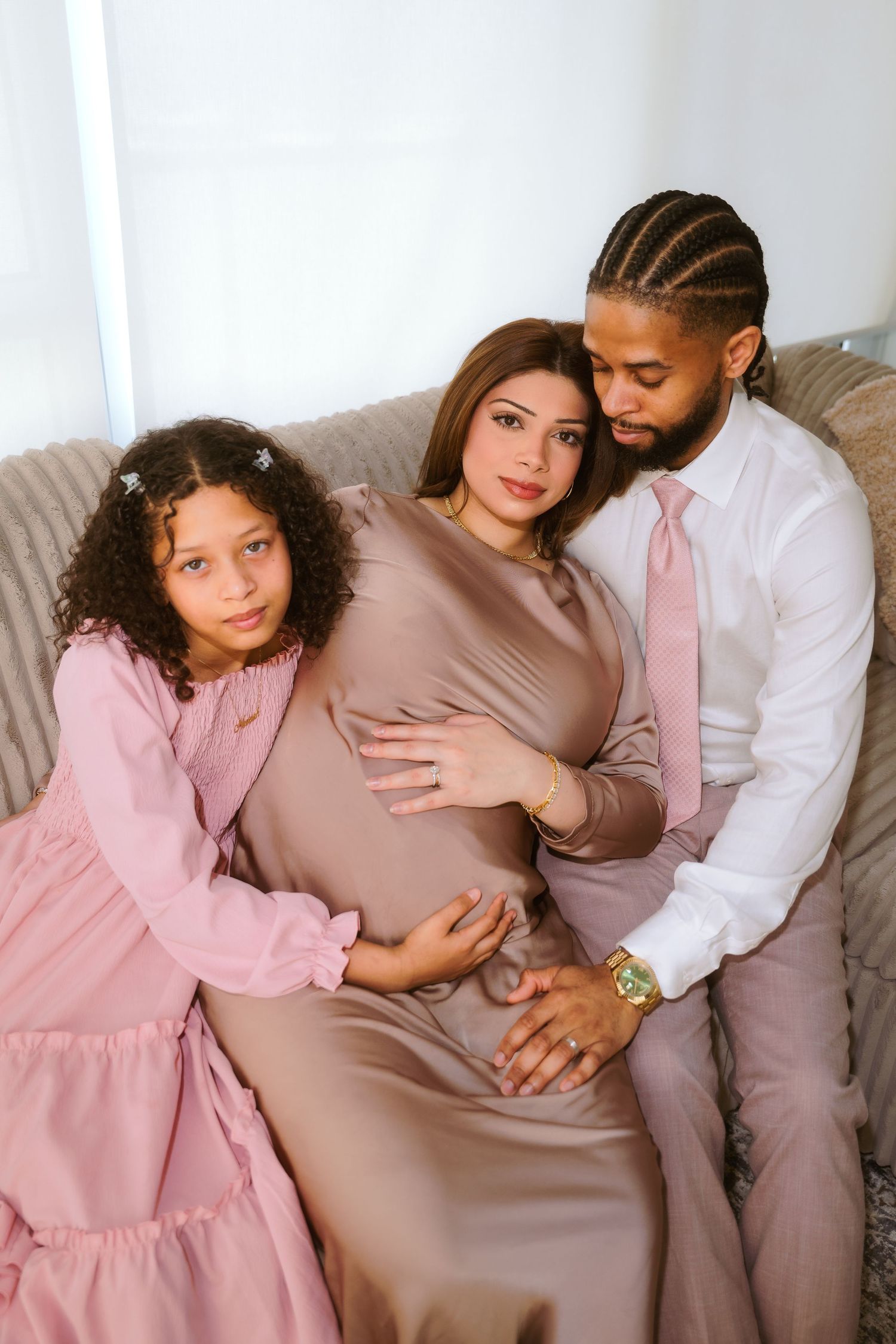 A family in coordinated pink and beige outfits sits together on a couch while expecting a new addition.