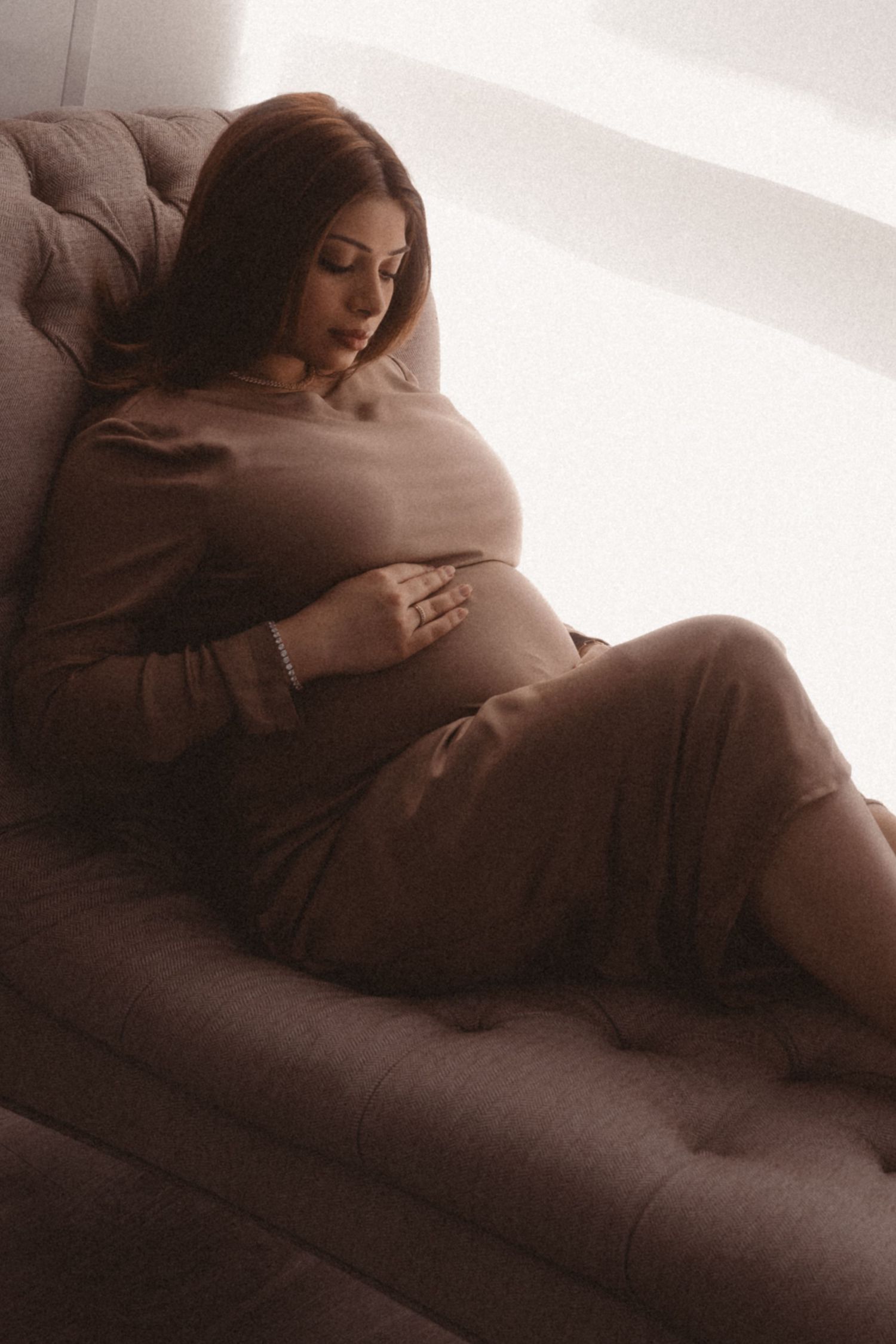 A pregnant woman in a brown dress rests peacefully on a tufted couch in soft, moody lighting.