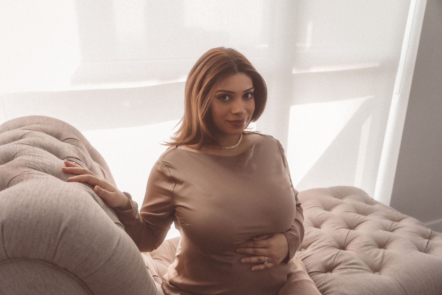 Woman in neutral toned clothing poses on sofa in soft window light during maternity photoshoot.