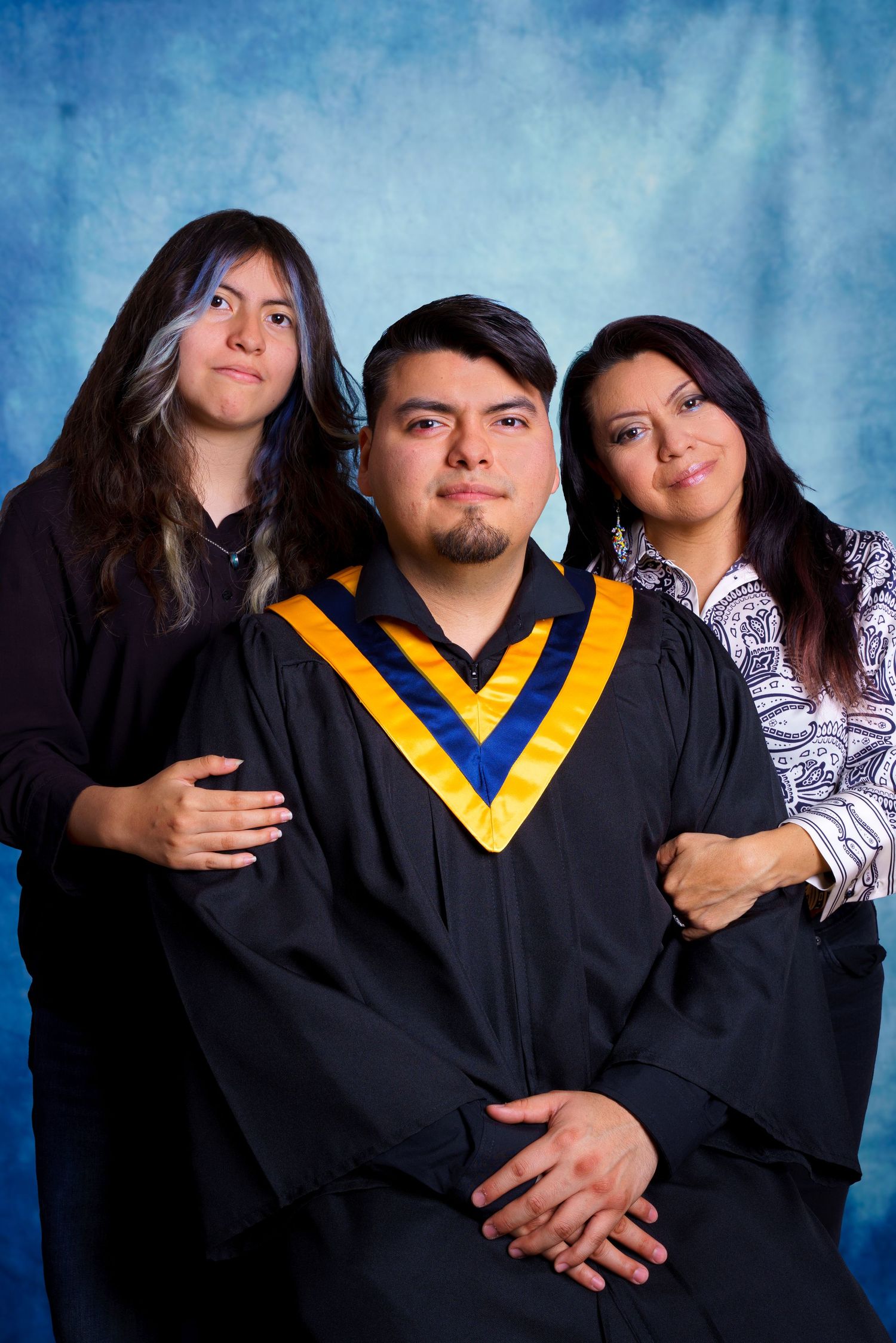 Family graduation portrait featuring graduate in black and gold regalia with two supporters against blue background.