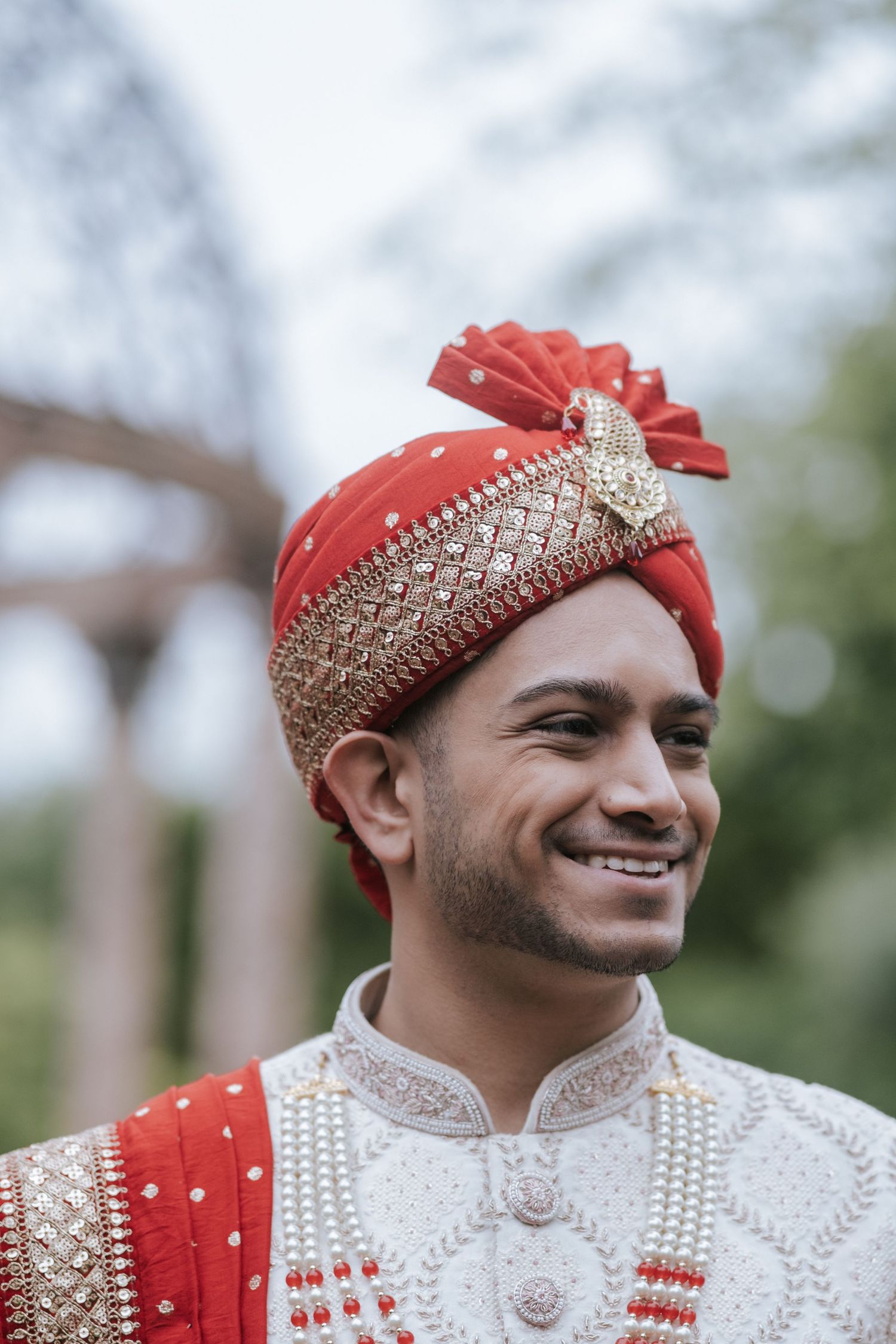 Handsome Indian groom smiling outdoors wearing a red and gold safa turban and white embroidered sherwani.