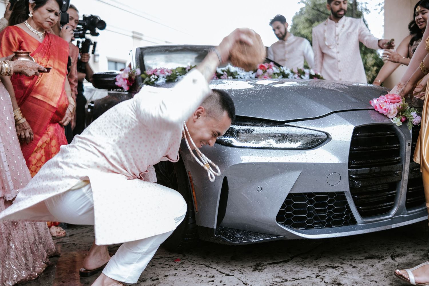 Groom in white kurta bows before flower-decorated BMW car during Indian wedding baraat tradition.