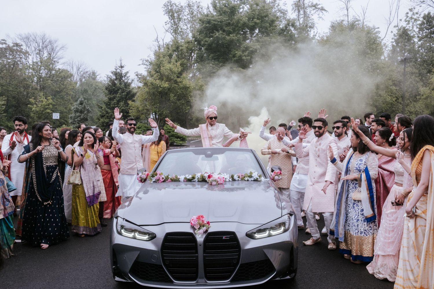 Indian groom stands in decorated BMW convertible surrounded by colorful wedding guests with smoke.