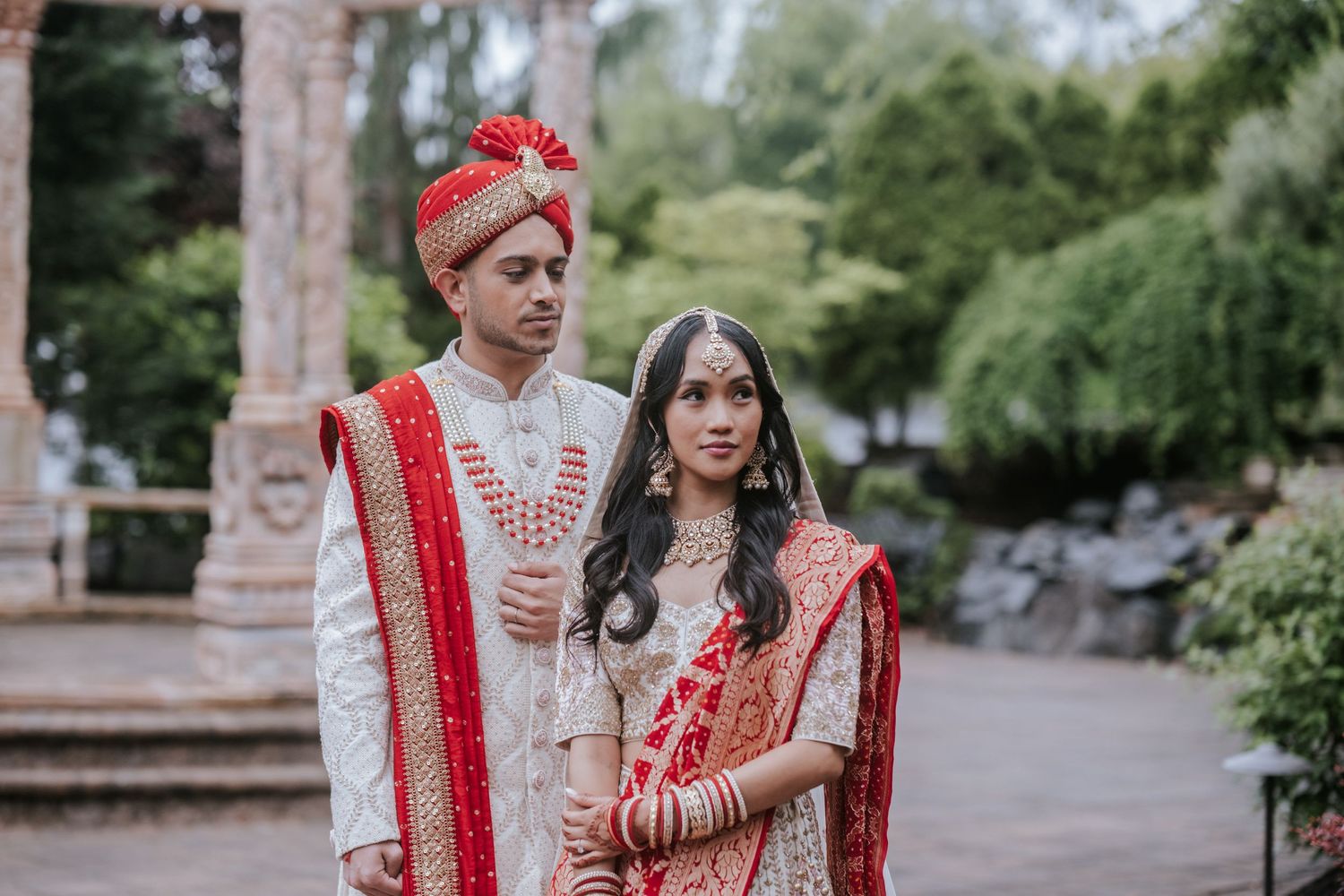 Indian bride and groom in traditional wedding attire standing together near stone pillars.