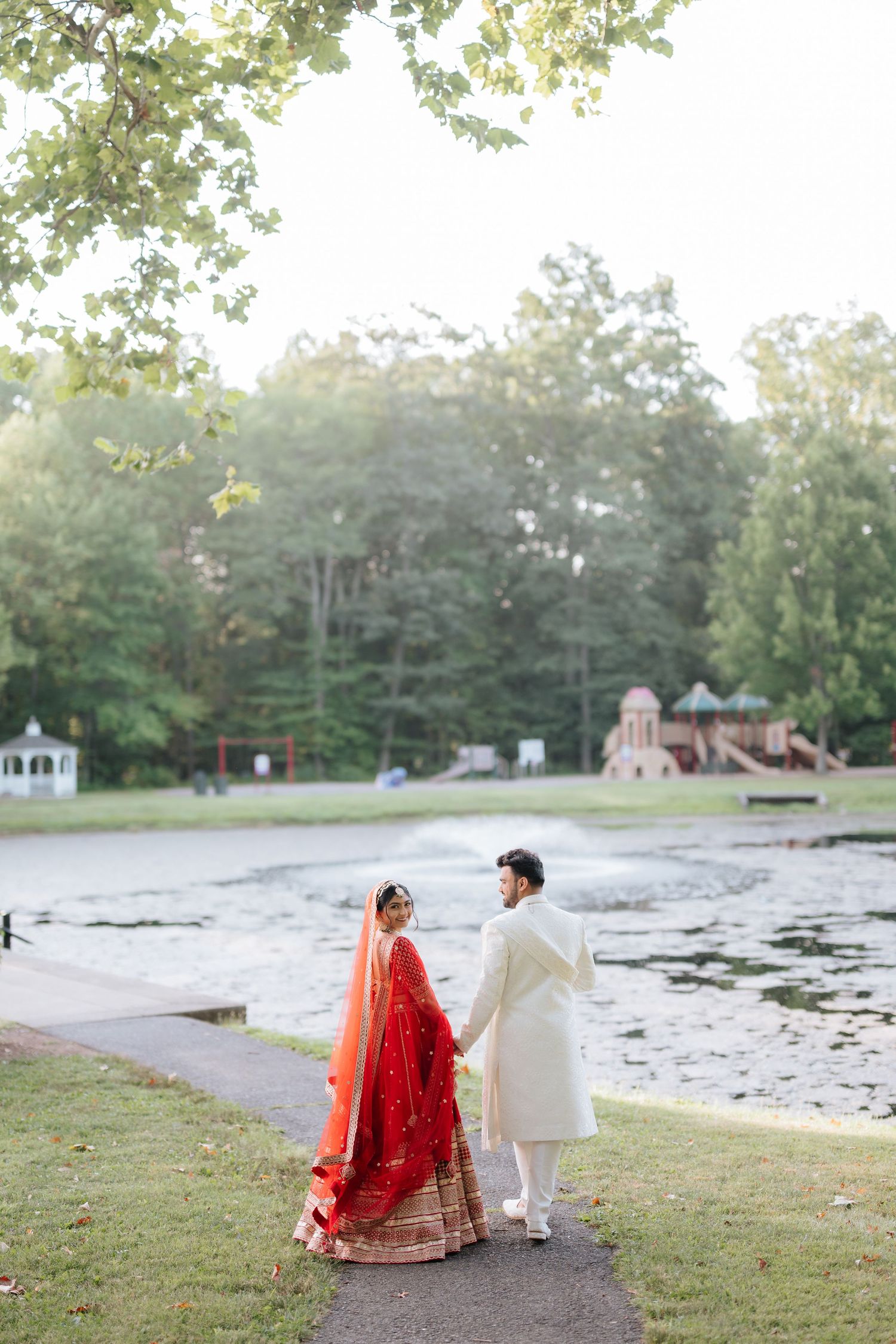 Indian wedding couple in red and white traditional attire holding hands by a serene lakeside path.