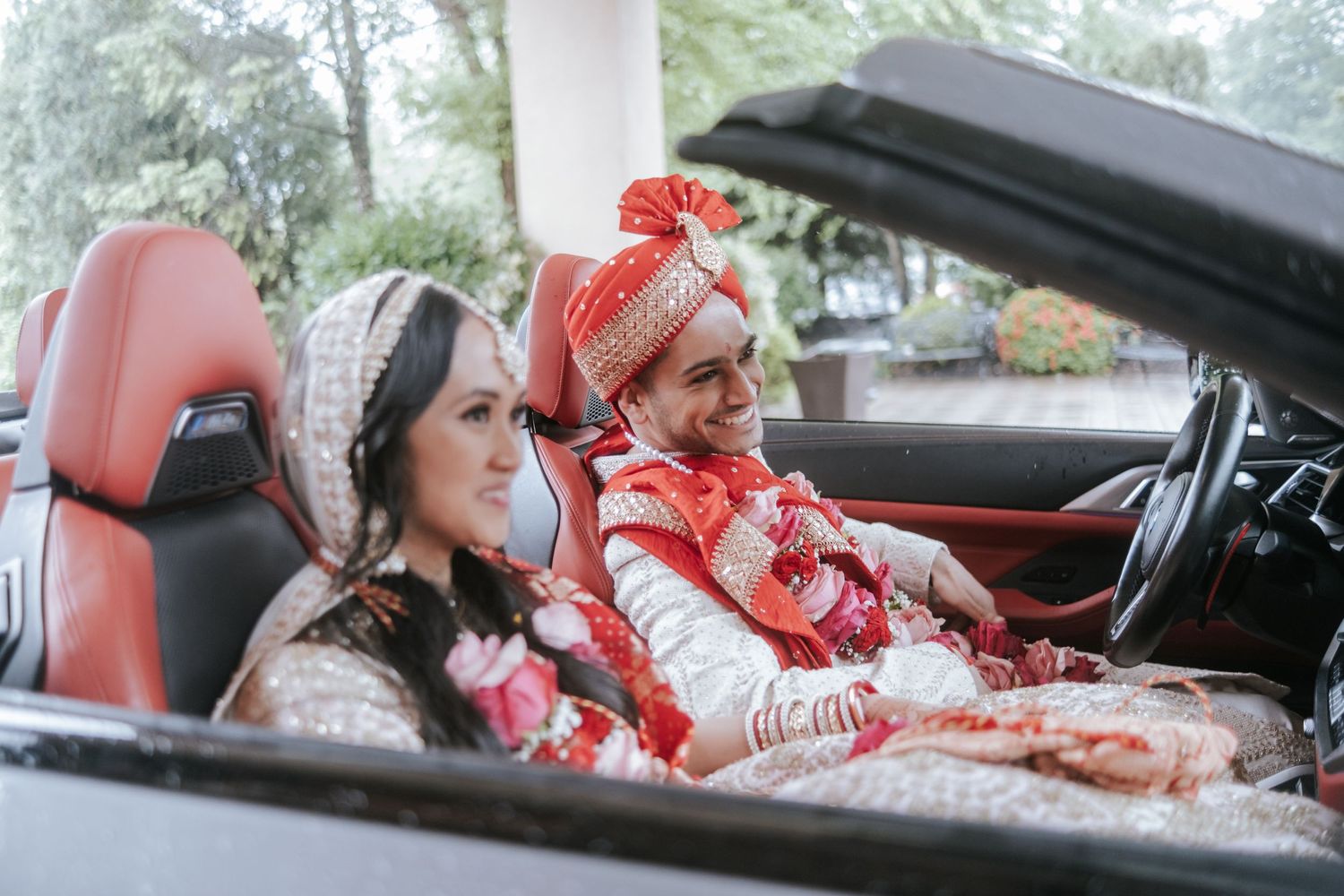 Indian bride and groom in traditional wedding attire sitting in a luxury convertible car.