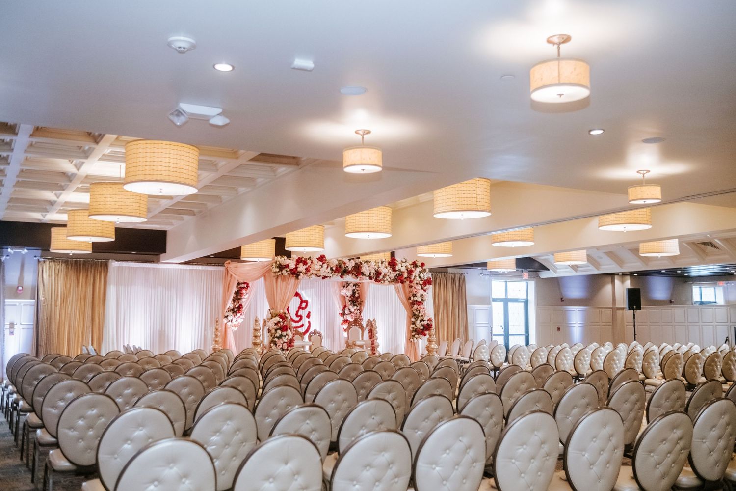 Spacious wedding ceremony venue with white tufted chairs and a floral mandap altar backdrop.