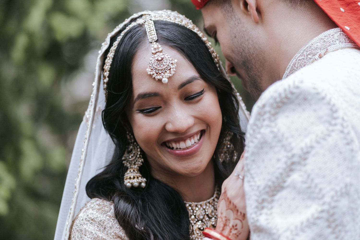 Smiling Indian bride in gold bridal lehenga and jeweled maang tikka shares intimate moment with groom at wedding.