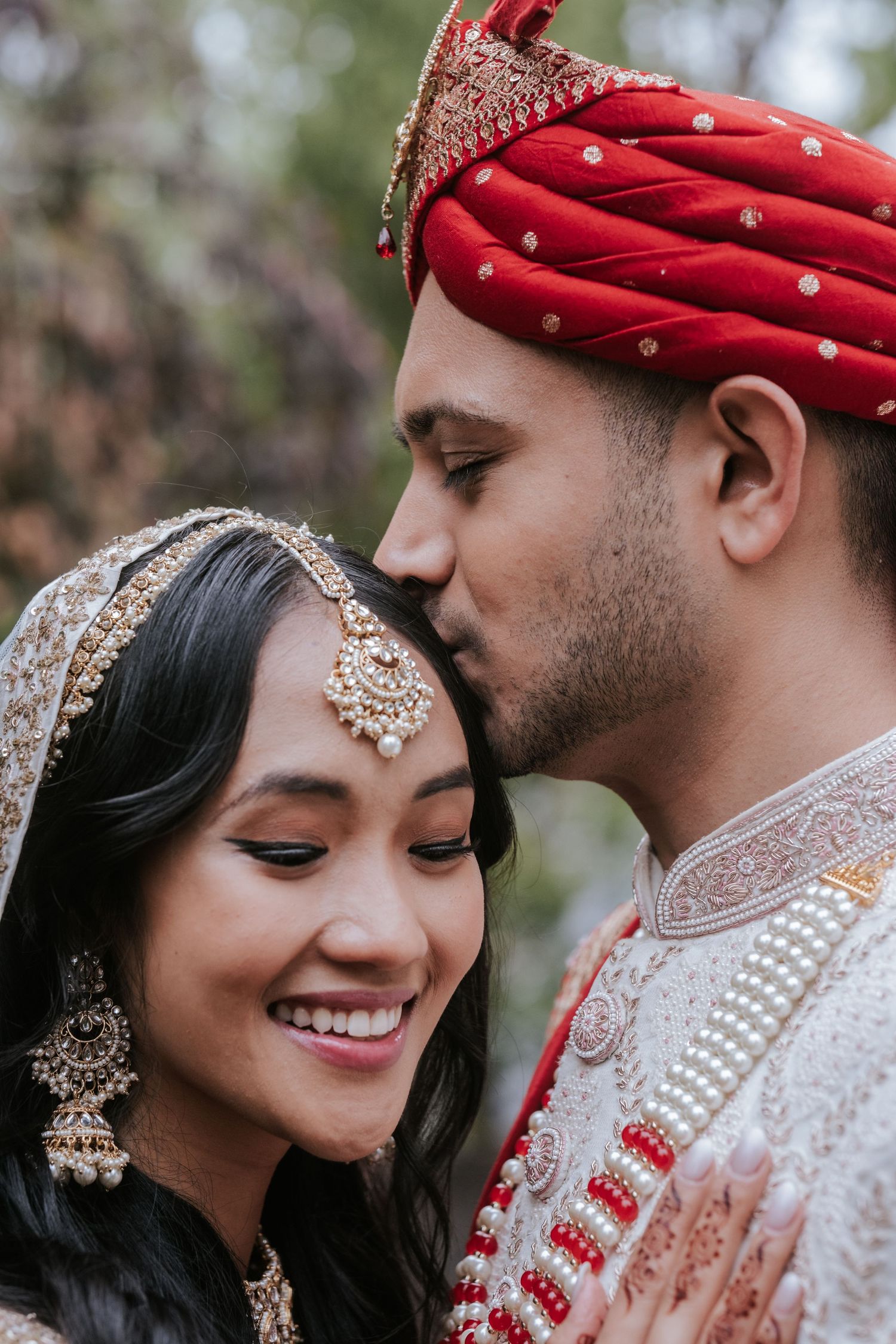 Indian groom in red turban gently kissing smiling bride's forehead during outdoor wedding photo session.