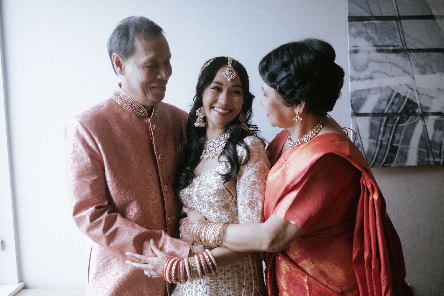 Indian bride smiles with parents in traditional wedding attire, lehenga and kurta.
