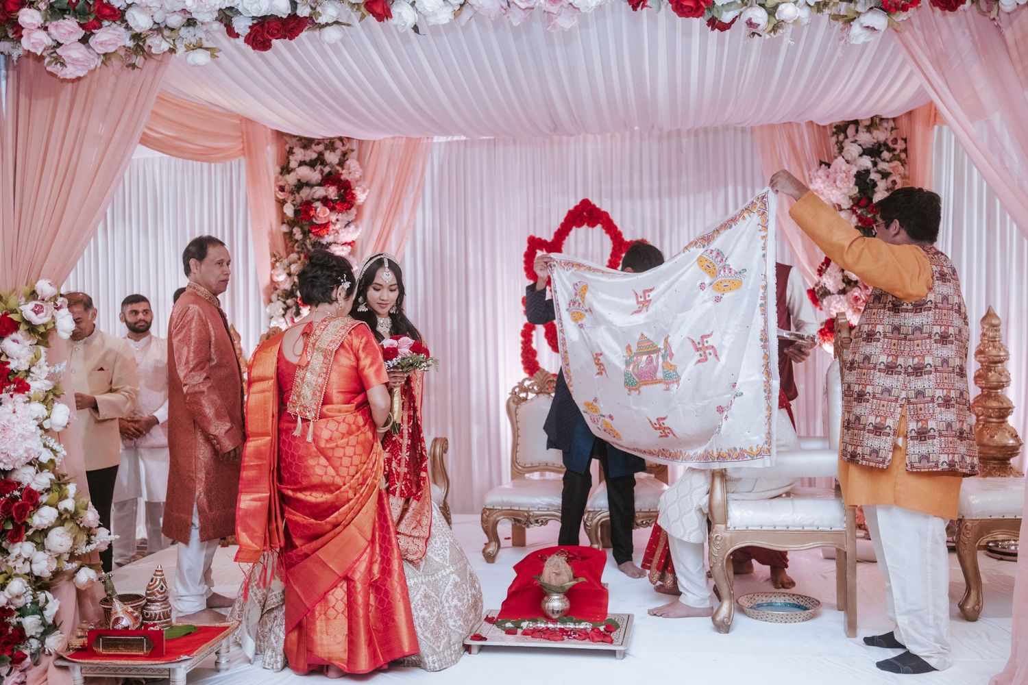 Hindu wedding ceremony at decorated mandap with red and white floral backdrop and Ganesh symbol.