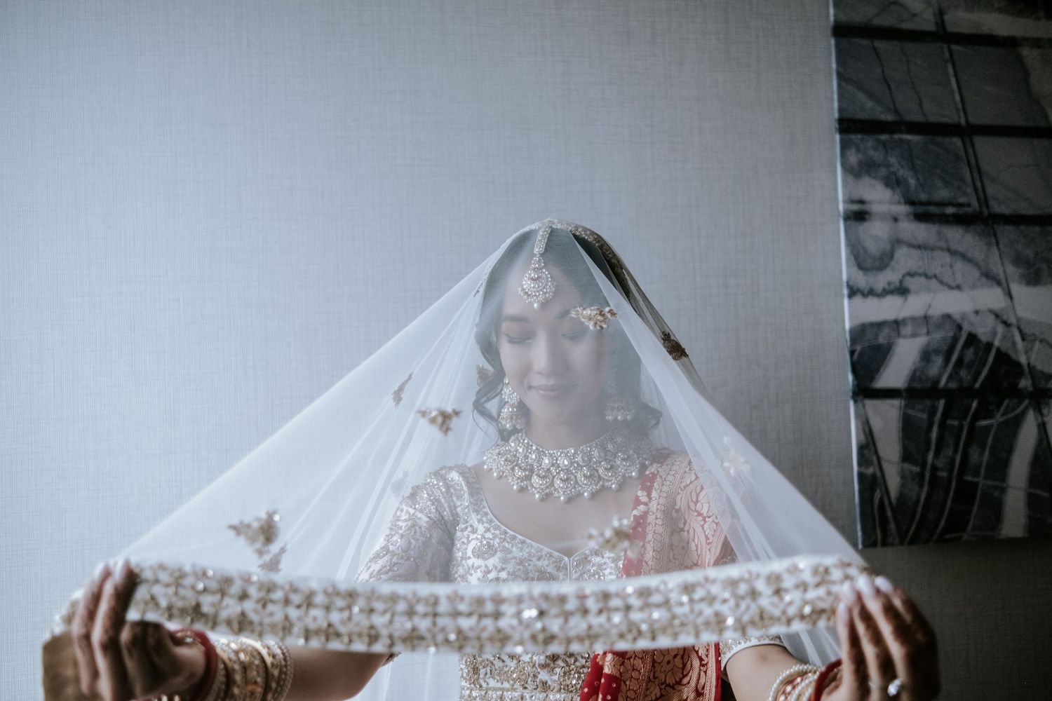 Indian bride holds embroidered veil over her face revealing intricate bridal jewelry.