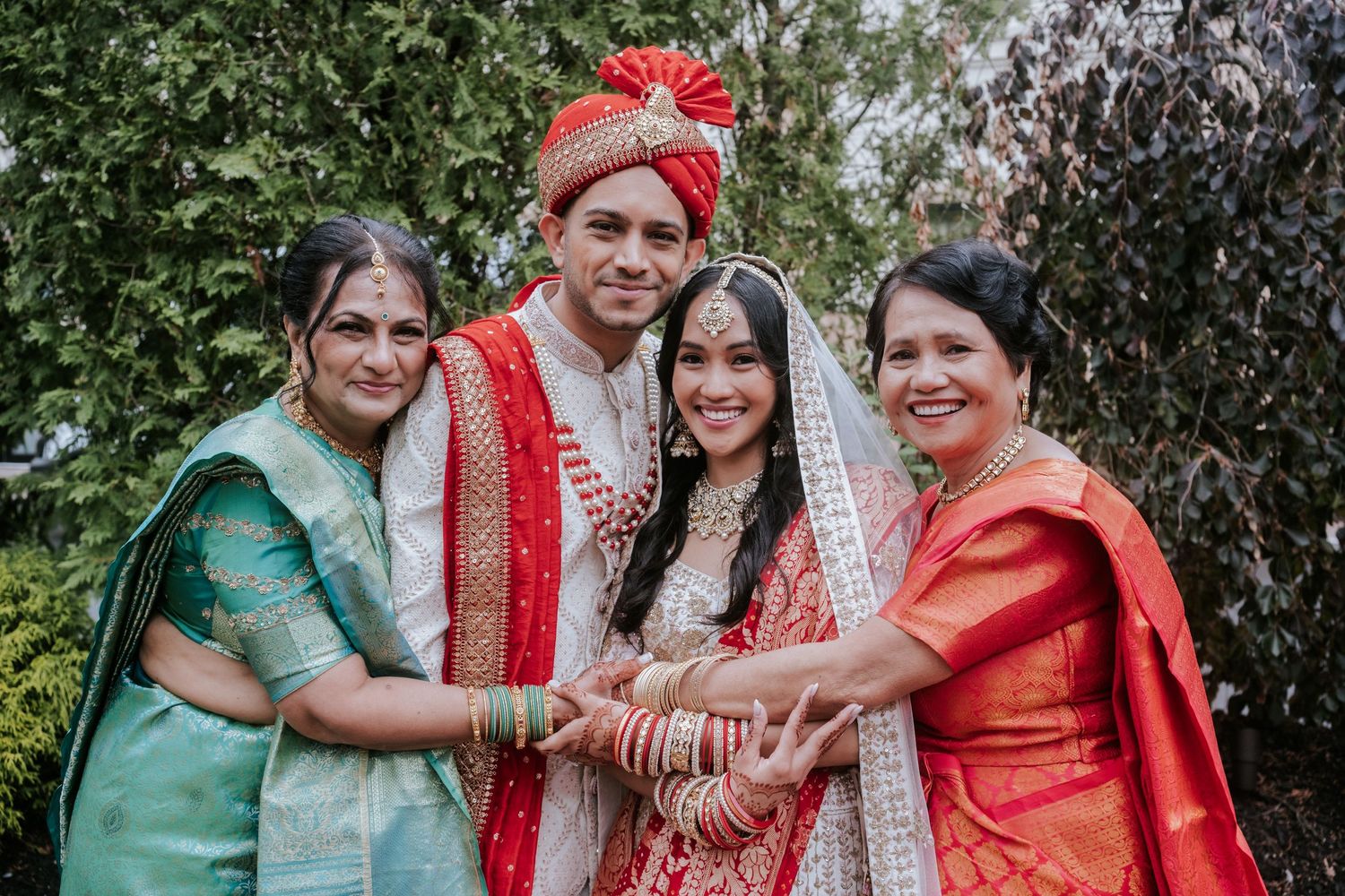 Indian bride and groom in traditional red wedding attire pose with two women in colorful saris outdoors.