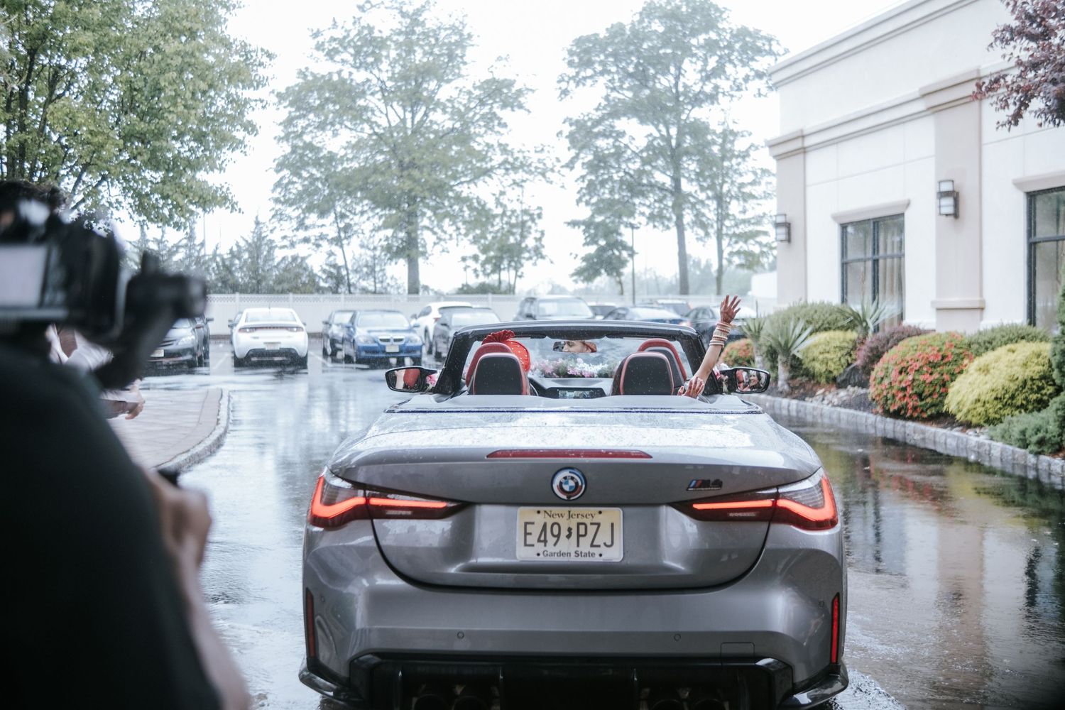 Photographer capturing Indian wedding couple in a gray BMW convertible on a rainy day.