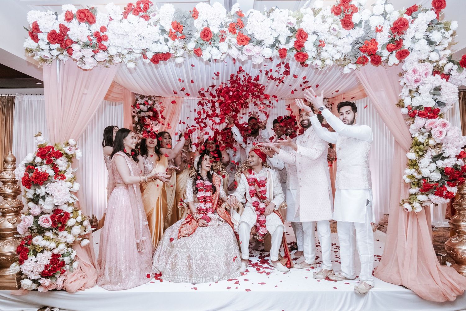 Bride and groom surrounded by family during colorful Indian wedding flower petal shower ceremony under floral mandap.