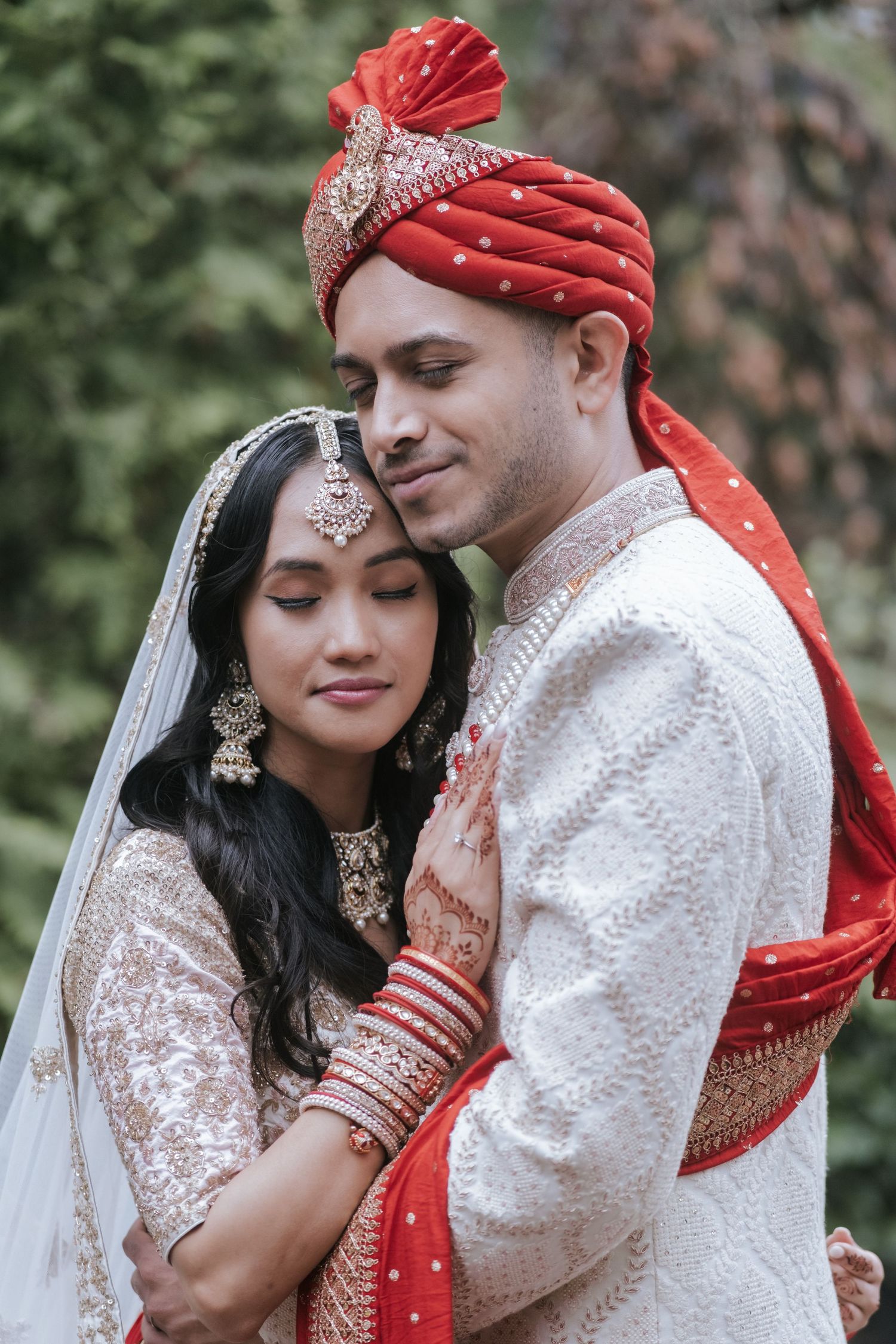 Indian bride and groom in traditional wedding attire share a tender embrace outdoors with lush greenery backdrop.