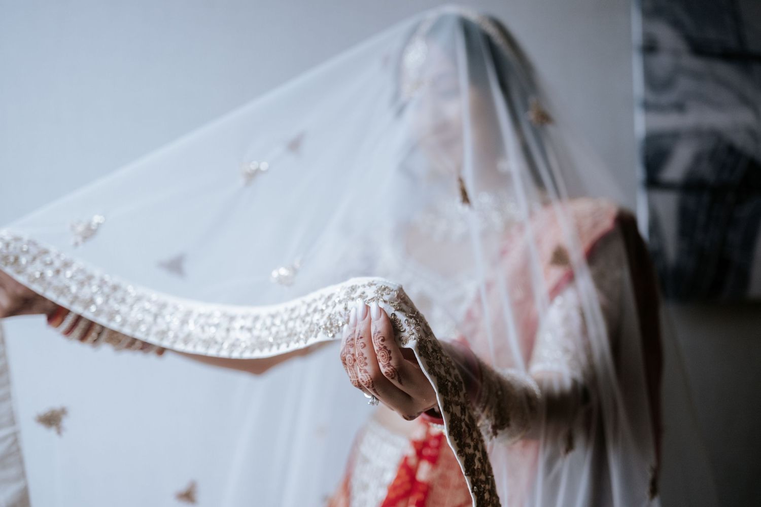 Indian bride lifting her white embroidered wedding veil with henna-adorned hands in traditional bridal attire.