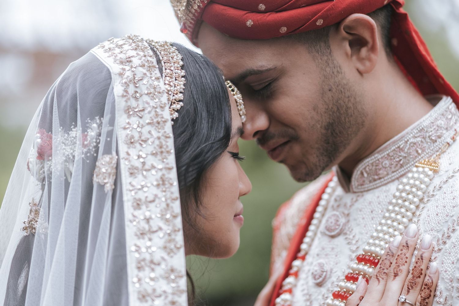 Indian bride and groom in ornate traditional wedding attire share an intimate forehead-to-forehead moment outdoors.