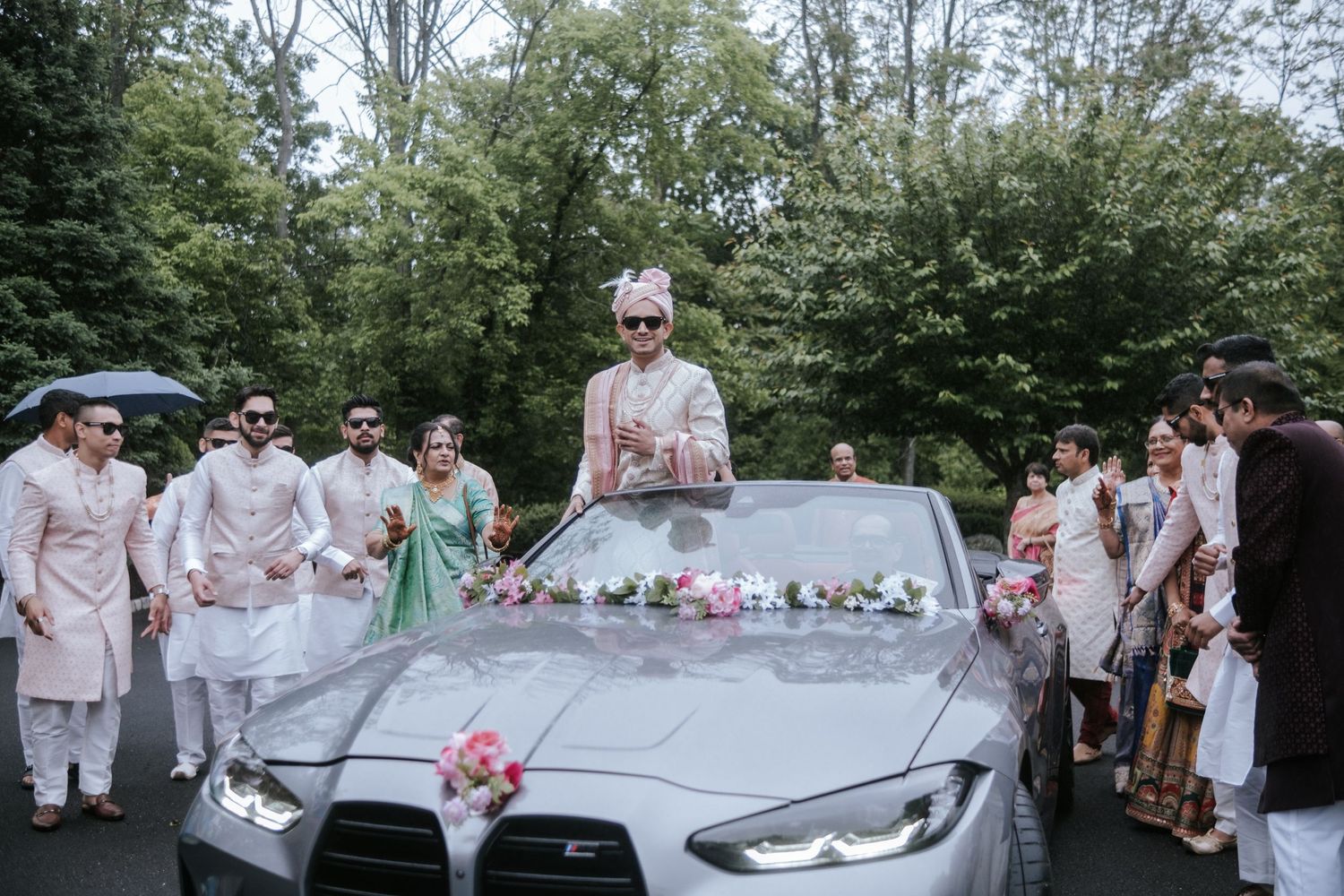 Groom in pink sherwani standing through sunroof of a flower-decorated car during Indian baraat procession.