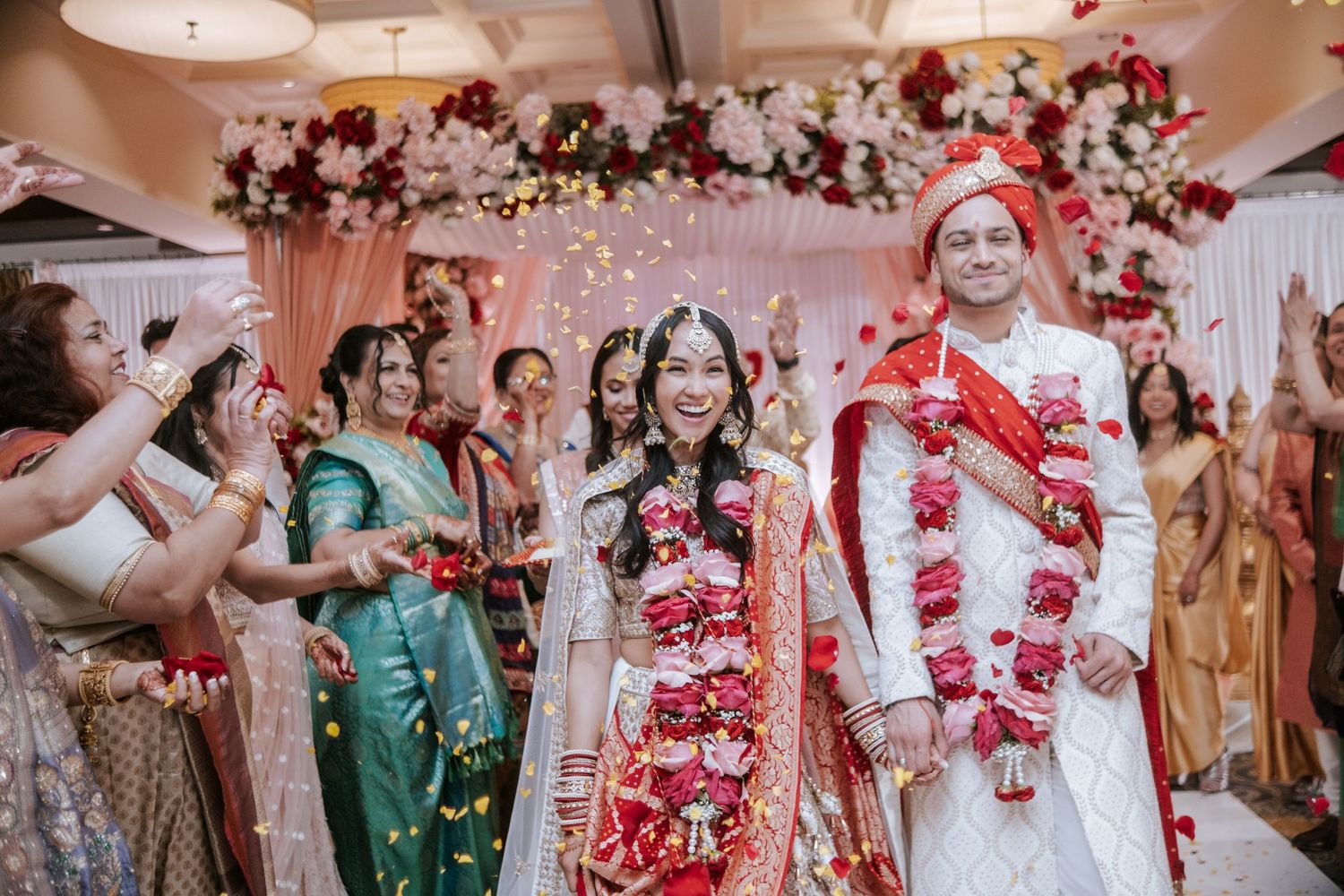 Joyful Indian wedding couple exits ceremony as guests toss colorful flower petals in celebration.