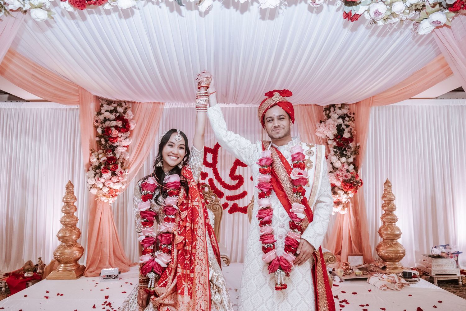 Indian bride and groom raising hands triumphantly in front of a decorated pink mandap after their wedding ceremony.