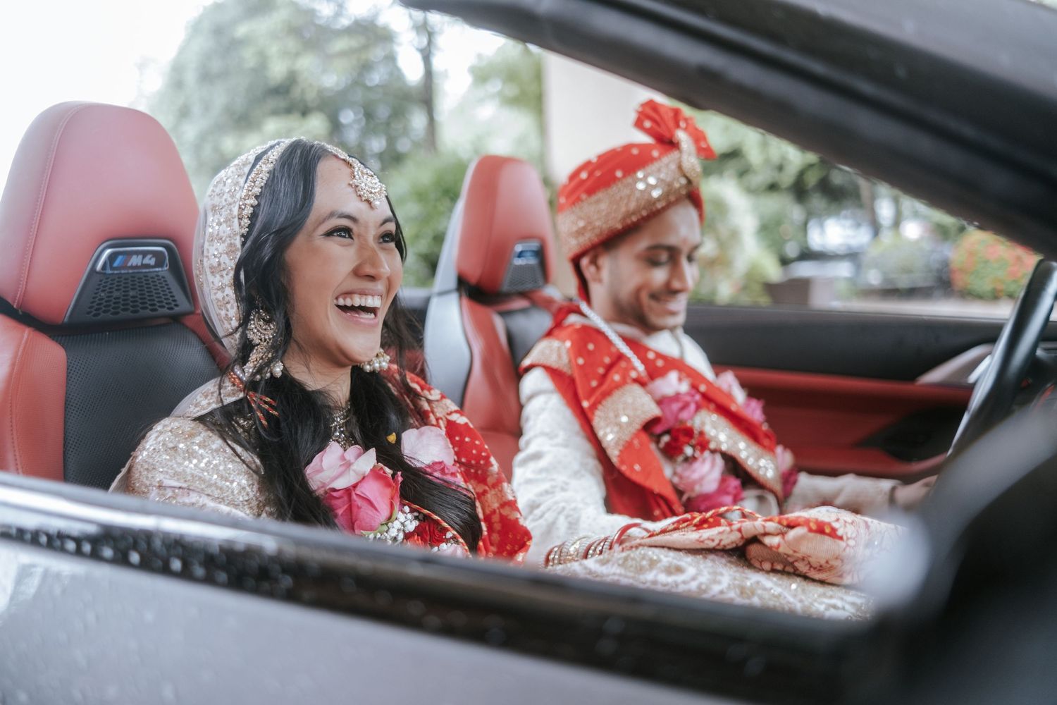 Smiling Indian bride and groom dressed in traditional wedding clothes seated in a red-interior convertible.