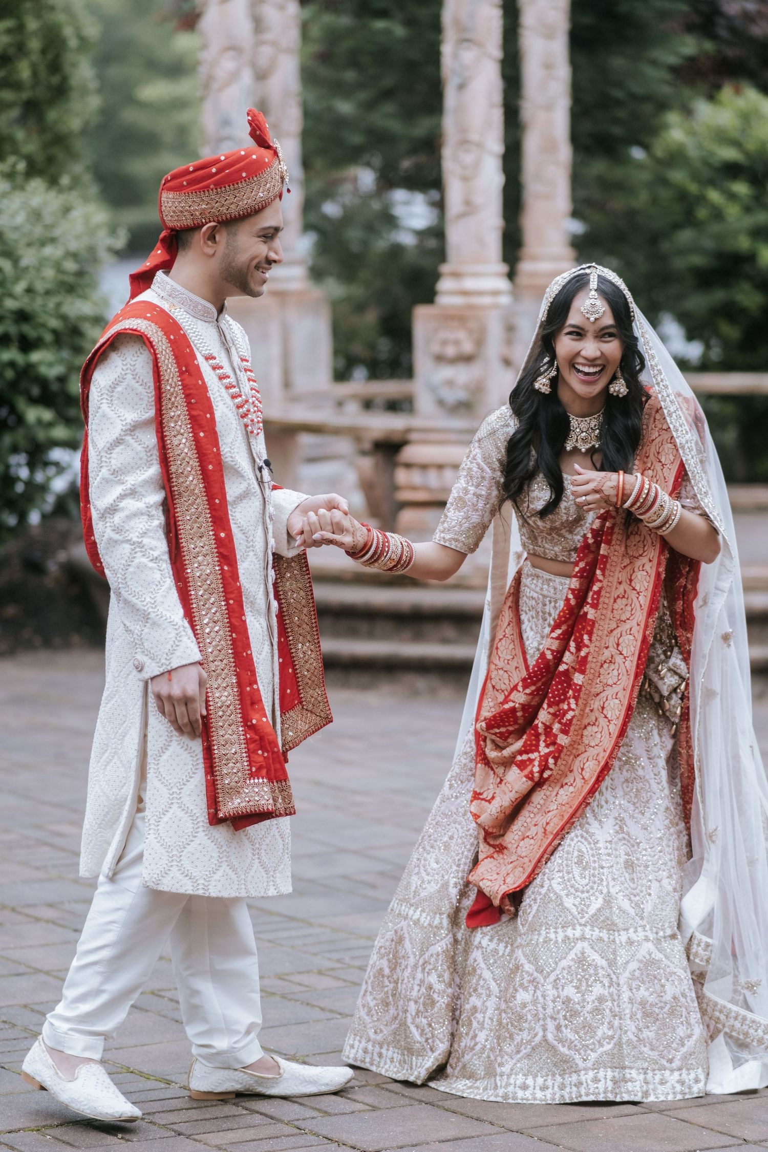 Indian bride and groom laughing together in matching white and red traditional wedding outfits outdoors.
