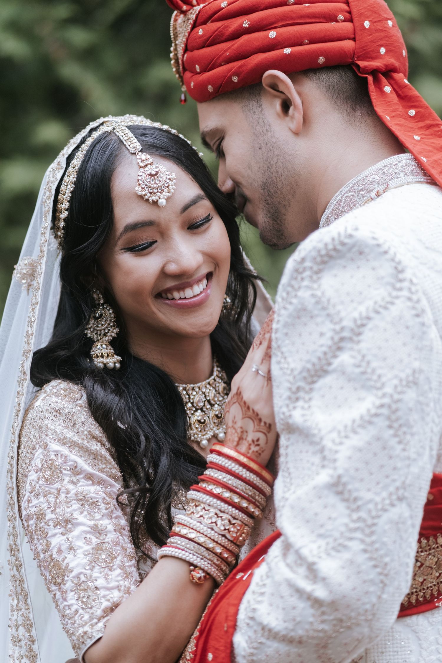Radiant Indian bride in white lehenga with mehndi and red bangles smiles joyfully beside groom in red turban.