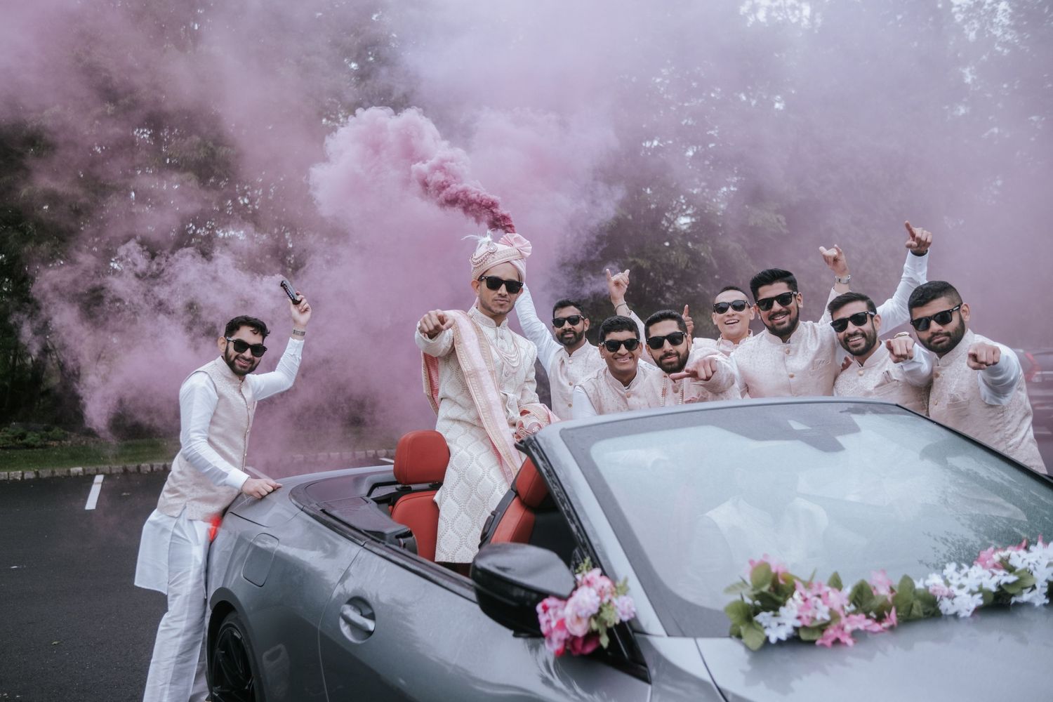 Groom and groomsmen in white suits celebrate with pink smoke bomb beside a decorated convertible car.