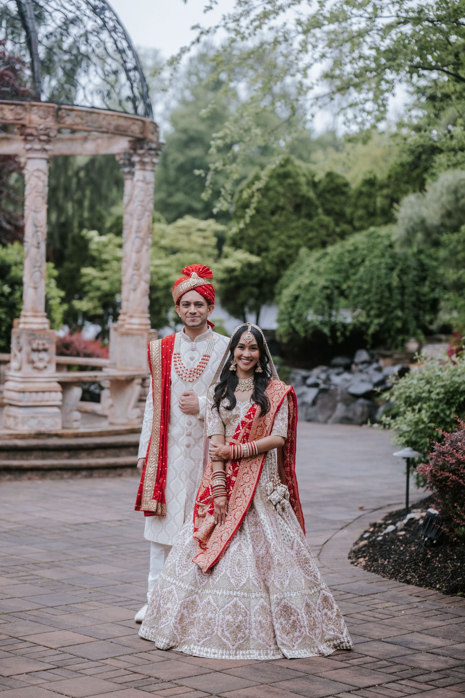 Indian bride and groom posing together in traditional red and white wedding attire outdoors.