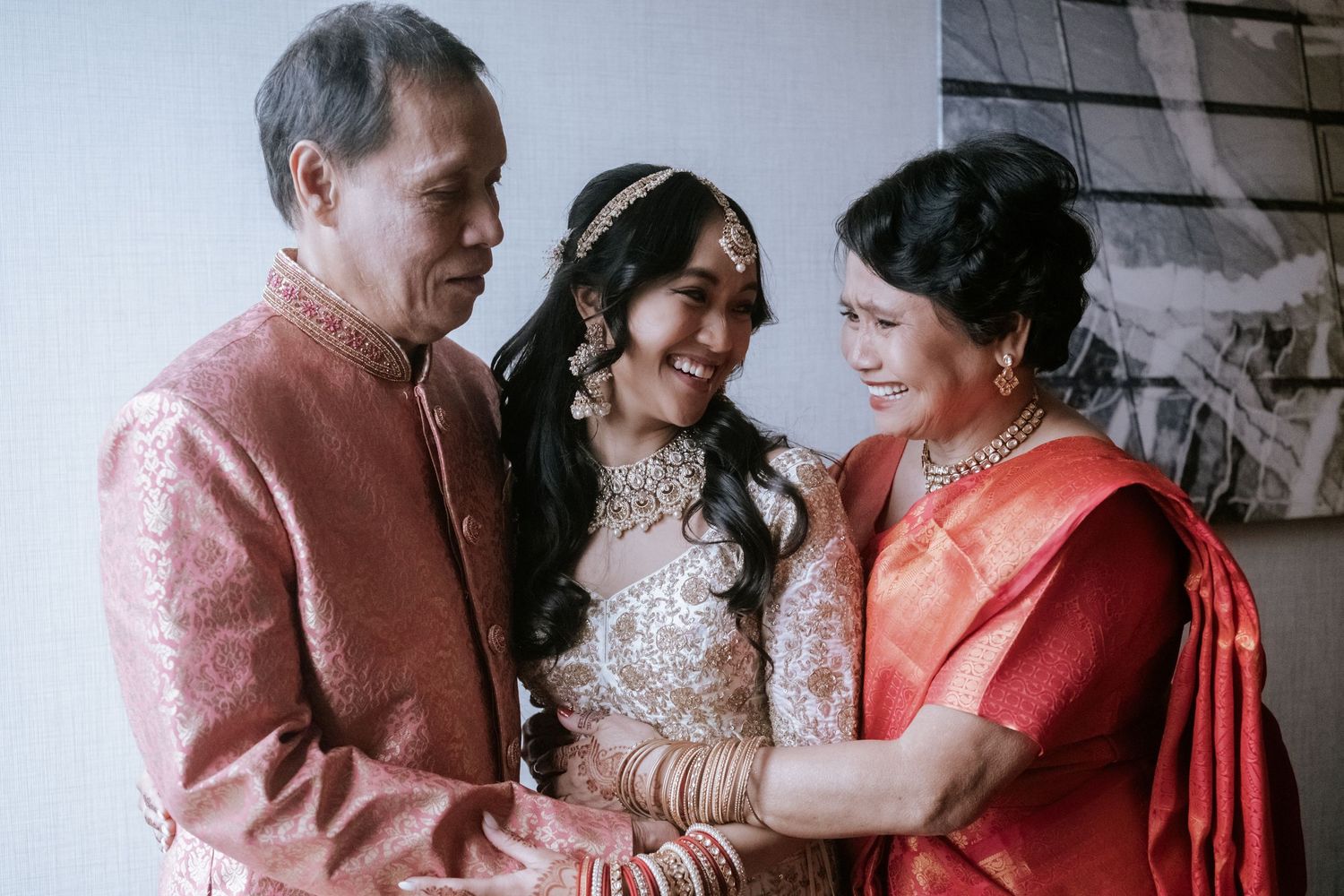 Indian bride in white lehenga embraces her parents wearing traditional red and gold wedding attire.
