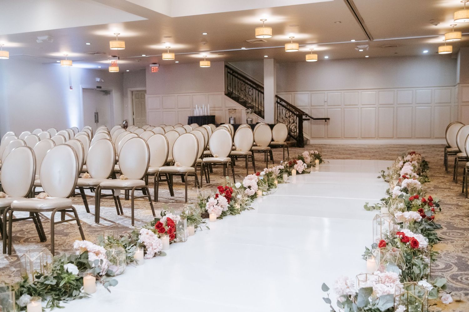 Elegant wedding ceremony aisle with white floral arrangements and cream chairs in a grand ballroom.